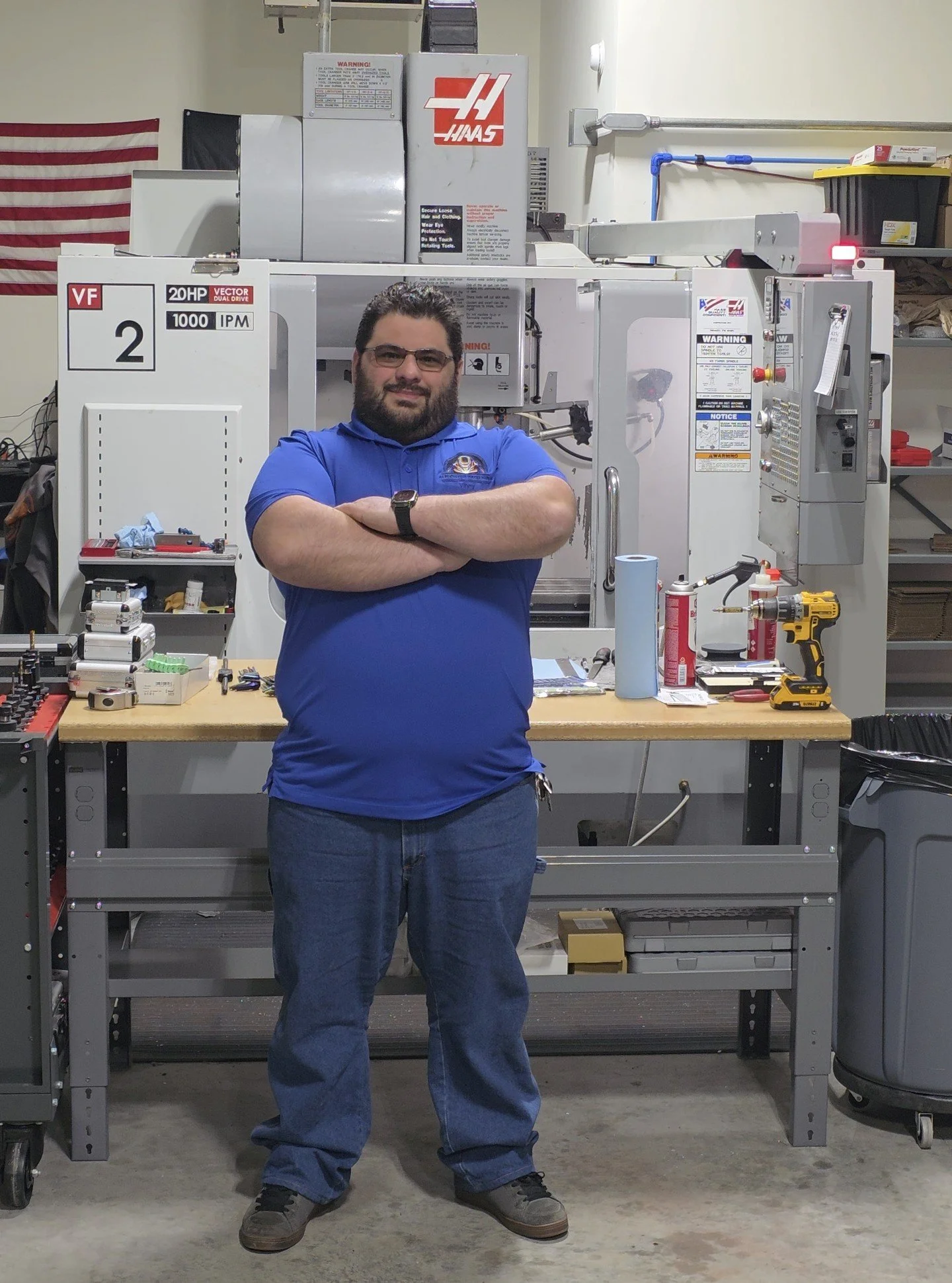 A man standing in front of a large industrial machine in a workshop, with arms crossed and smiling. The machine has various warning and instruction labels, tools, and equipment on the workbench in front of him.