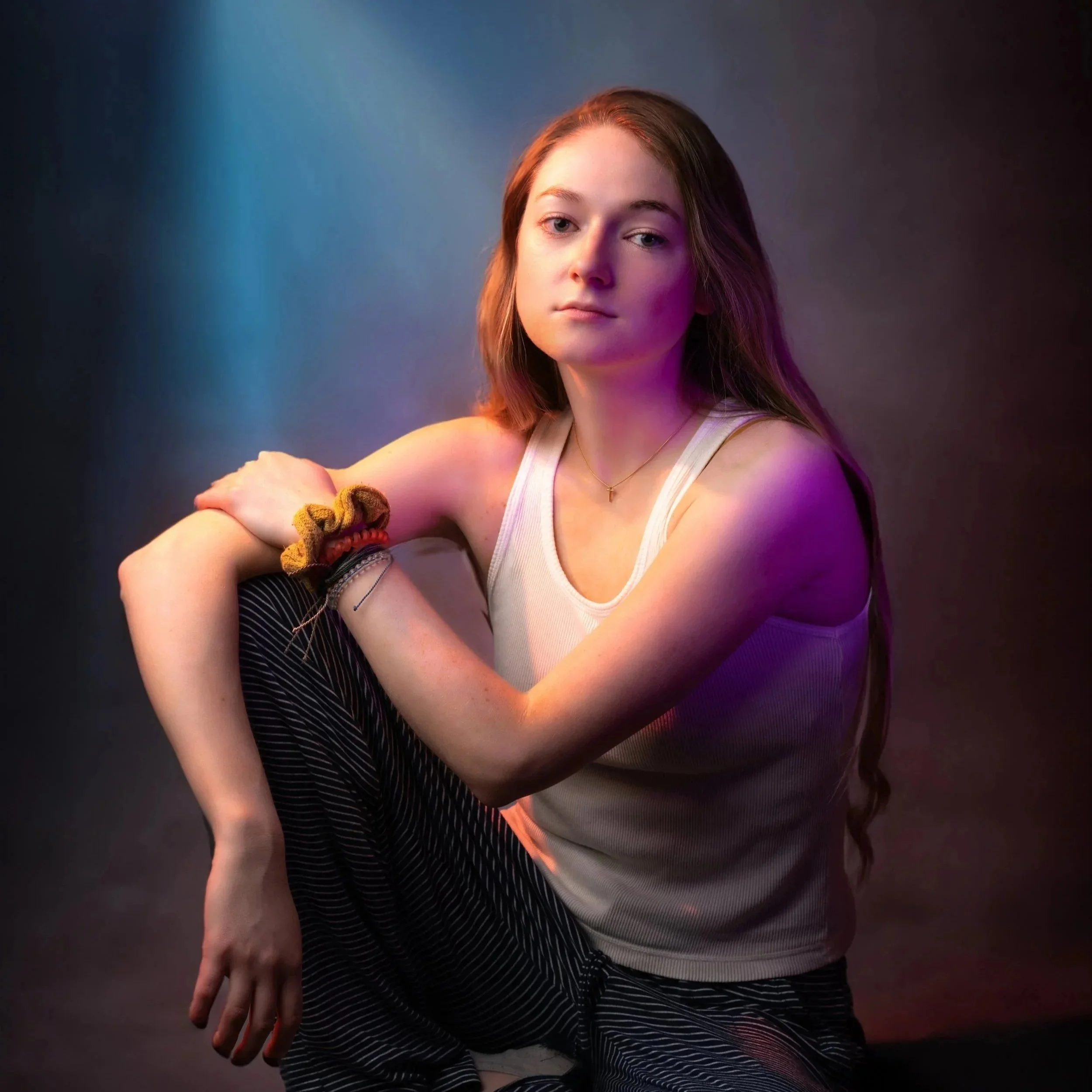 A young woman with long brown hair wearing a white tank top and striped pants, sitting in front of a dark background illuminated by multicolored lights, looking at the camera with a neutral expression, with her arms resting on her knee.