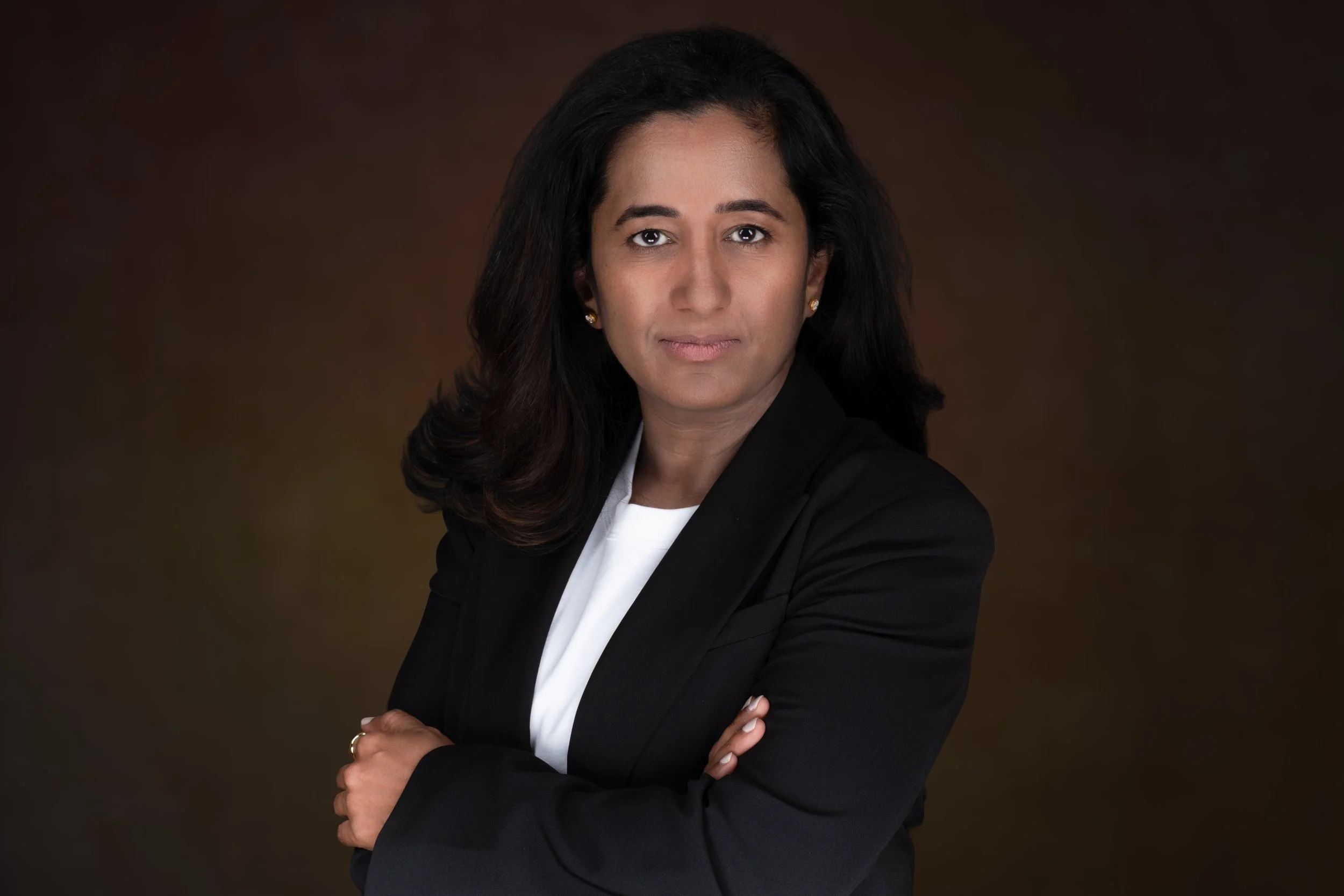 Professional portrait of a woman with dark hair, wearing a black blazer and white top, with arms crossed against a dark brown background.