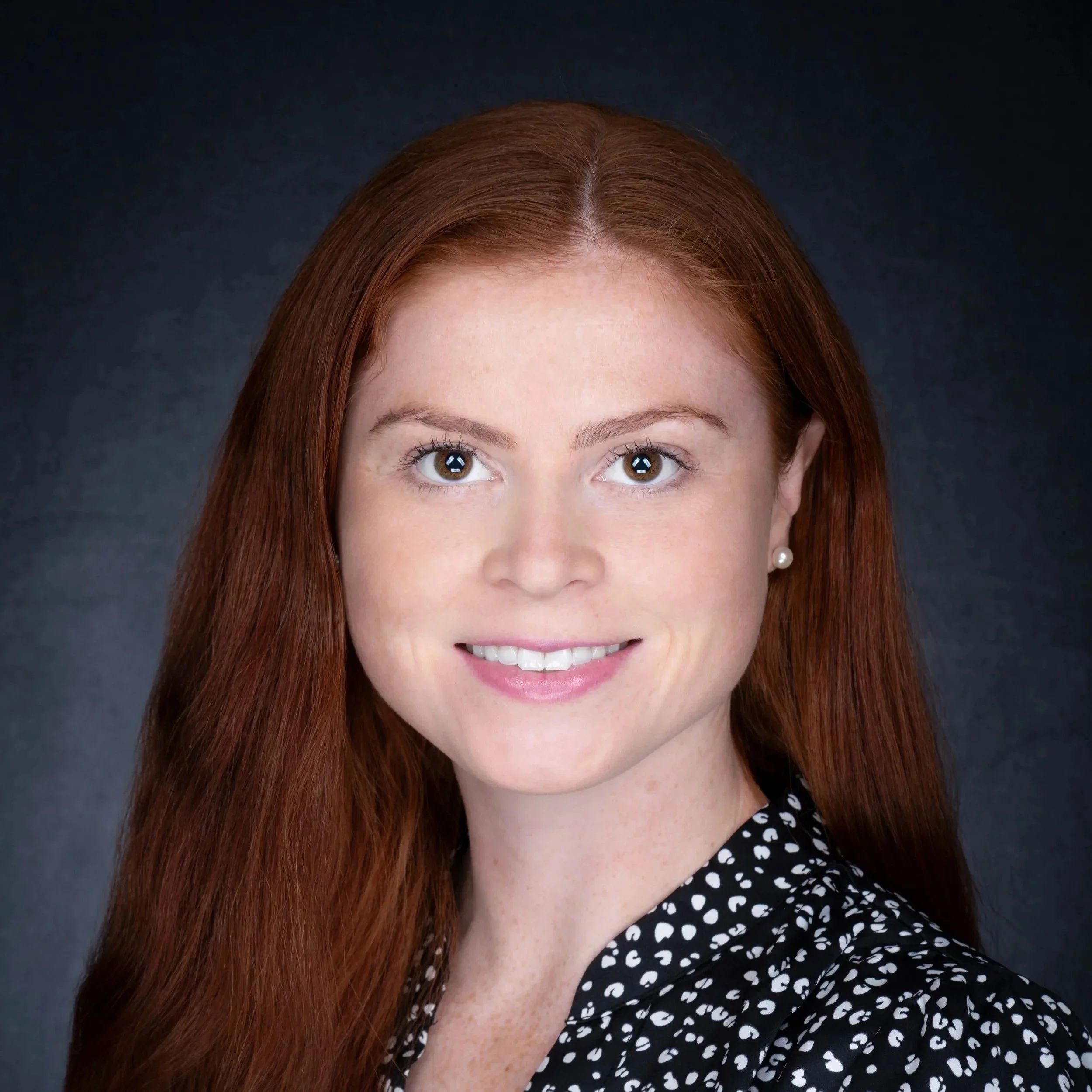 Headshot of a woman with red hair, wearing a black and white polka dot top, smiling against a dark background.