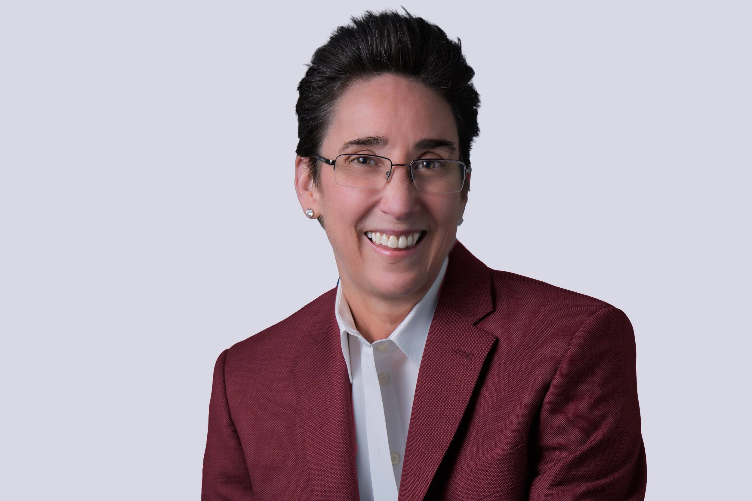 A woman with short dark hair, glasses, and earrings, smiling while wearing a burgundy blazer and white shirt against a light gray background.