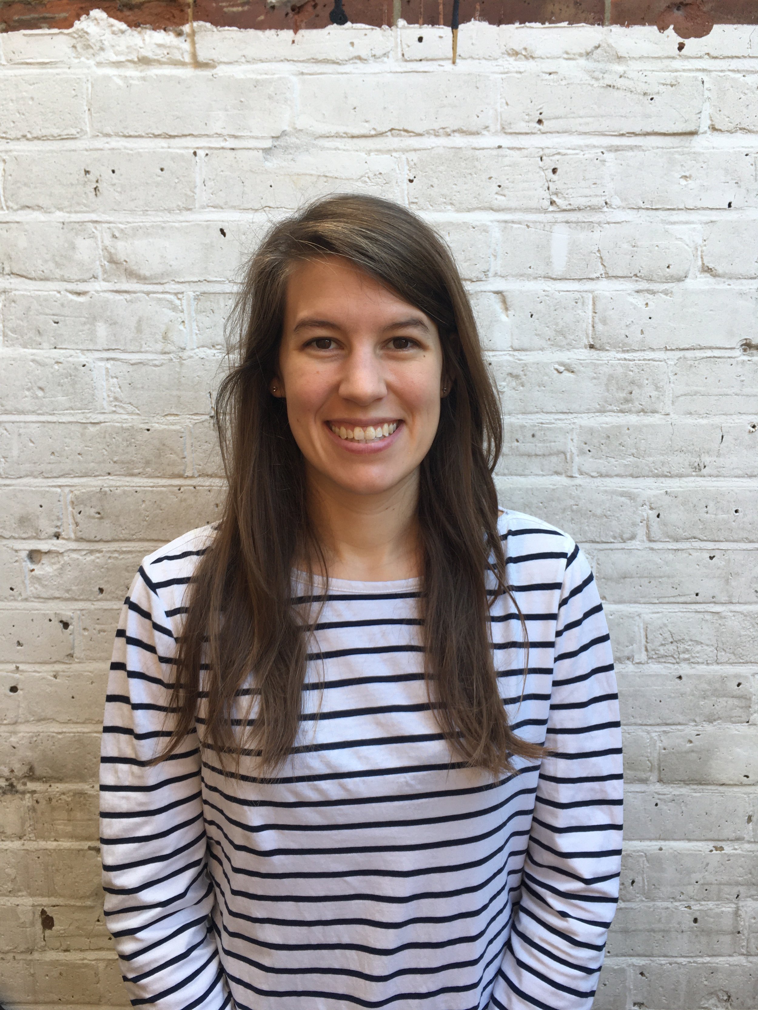 A young woman with long brown hair and a striped shirt standing in front of a white brick wall.
