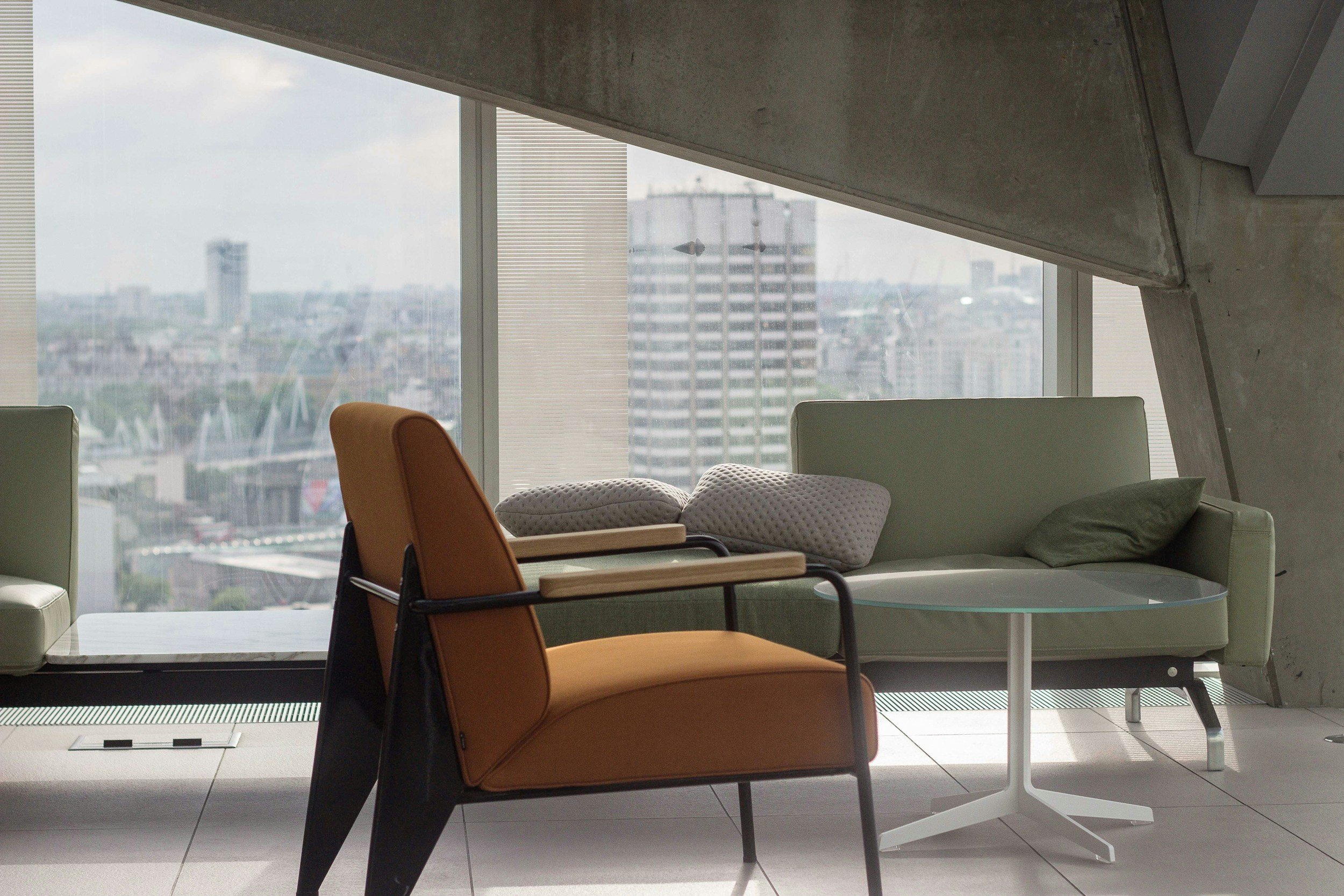 Interior of a modern office with a green sofa, two pillows, an orange chair, and a glass-top coffee table against large windows showing a cityscape with high-rise buildings.