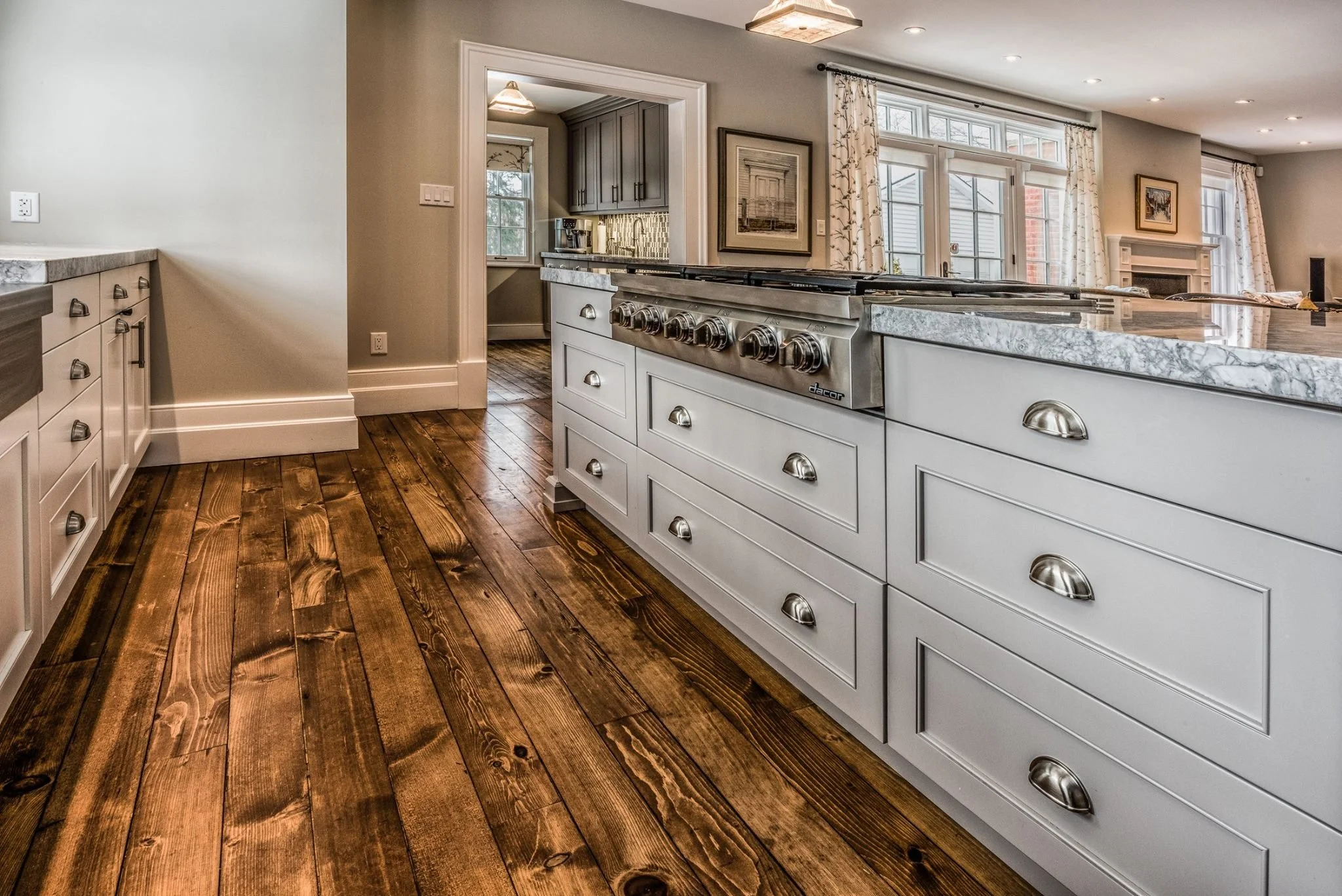 Interior view of a kitchen with white cabinets, a marble countertop, and a stainless steel stove. The kitchen opens into a living area with large windows and curtains, and hardwood floors.