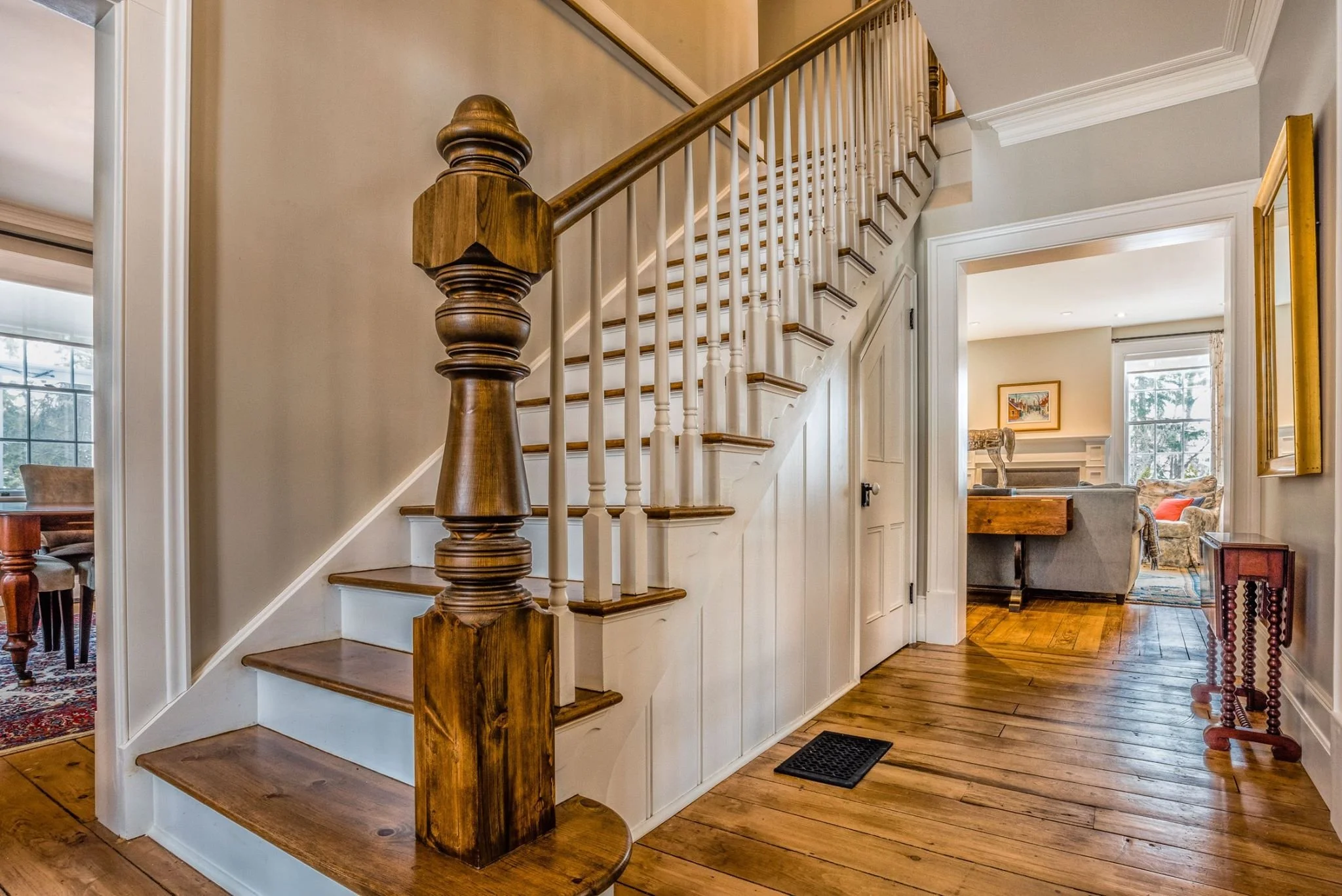 Interior view of a house with a staircase featuring wooden handrails and white spindles leading upstairs. The hallway has hardwood floors and is decorated with framed artwork and a mirror. Part of a living room with a fireplace and large windows is visible in the background.