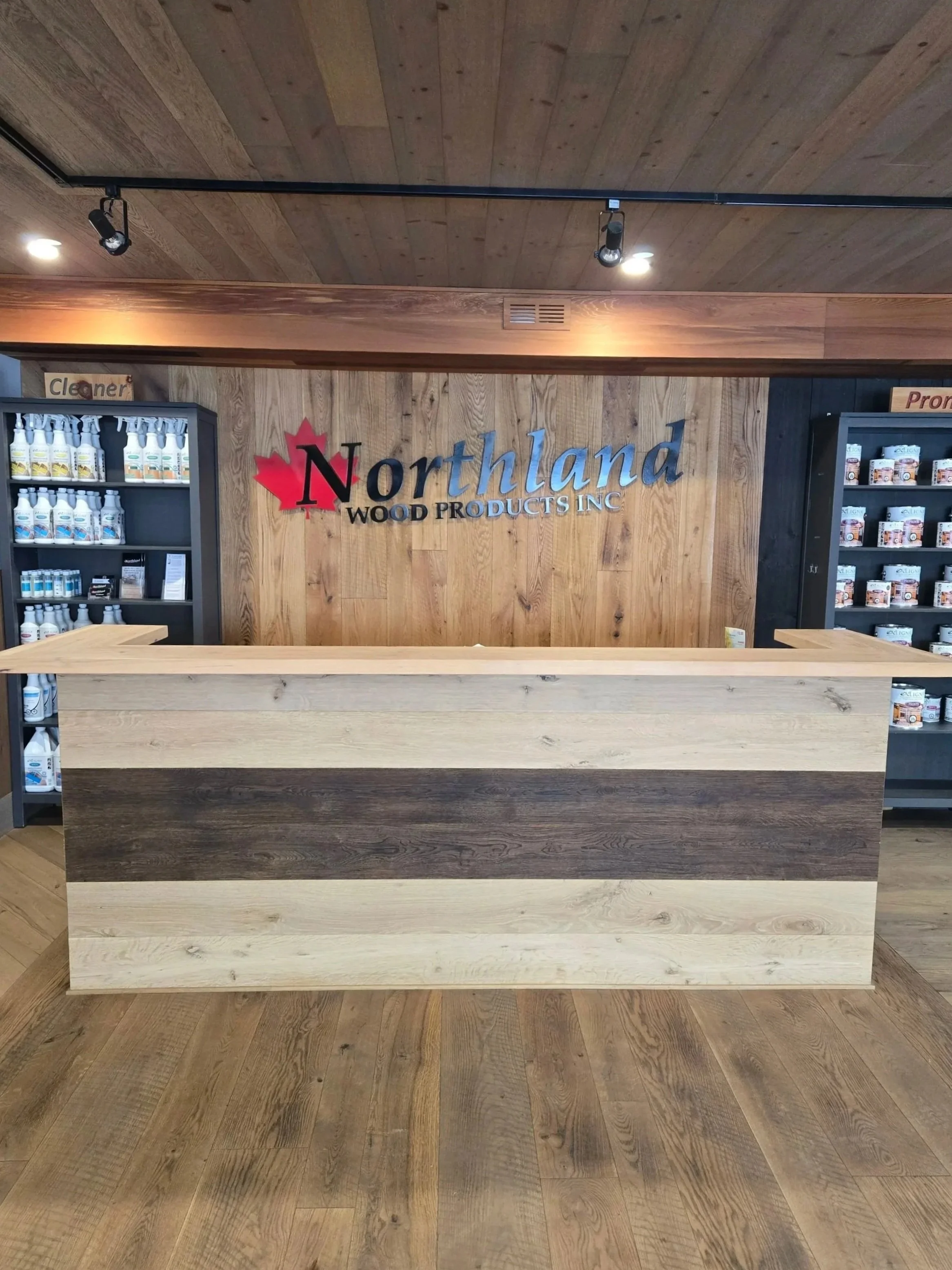 View of a wood counter and wooden wall with the sign 'Northland Wood Products Inc' and shelves with various bottles and jars of wood finishing products in the background.