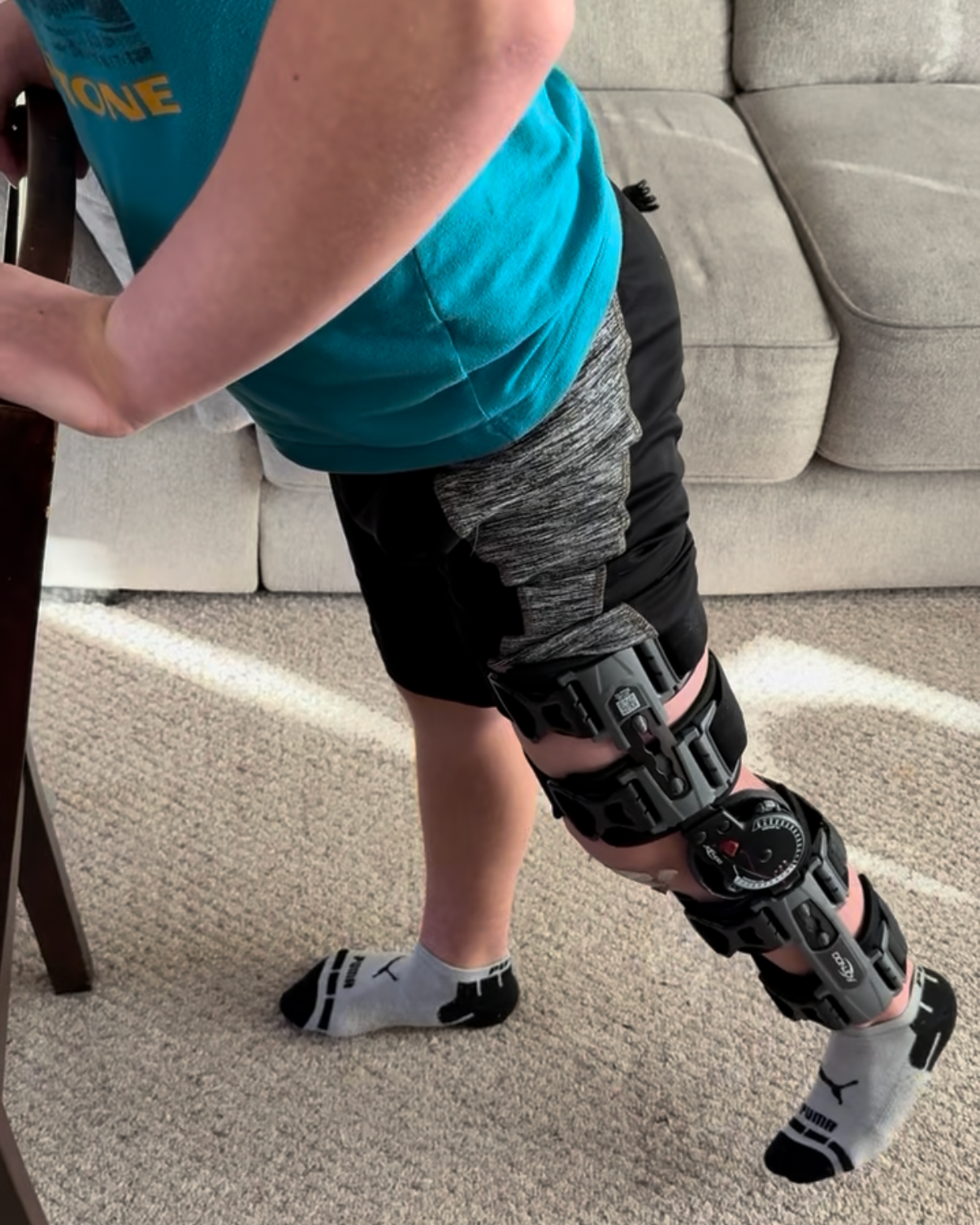 A person standing with one leg propped on a chair, wearing a knee brace or brace with a mechanical or orthopedic device, in a living room setting.