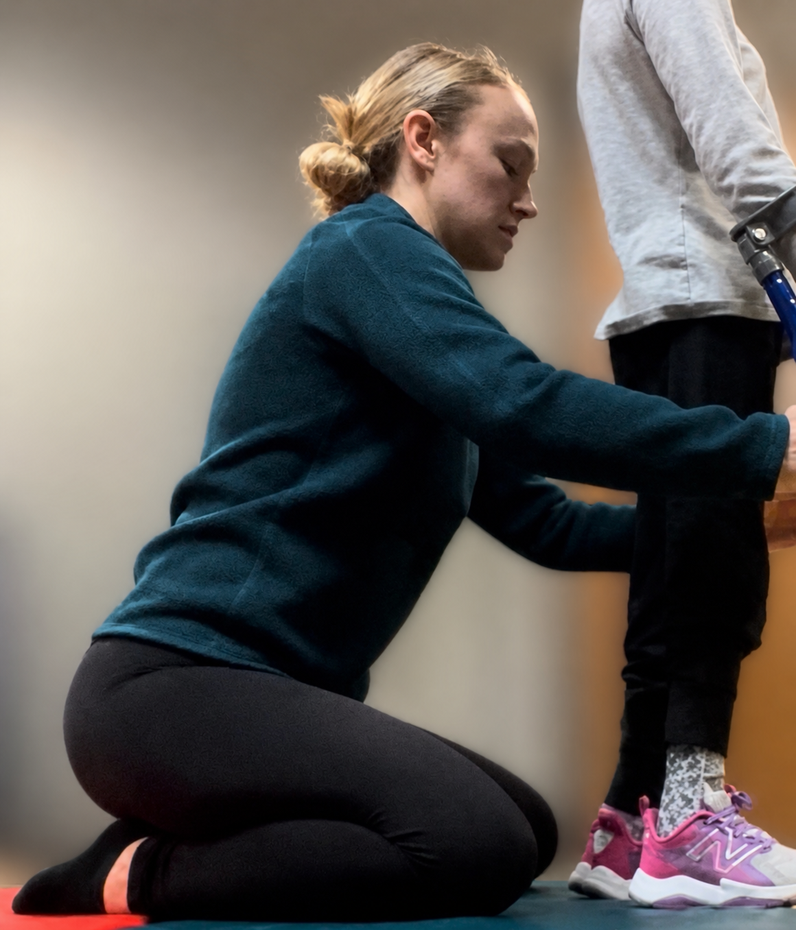 A woman in a teal sweater and black leggings is kneeling on the floor, assisting a girl in gray sweatshirt and black pants with pink sneakers.