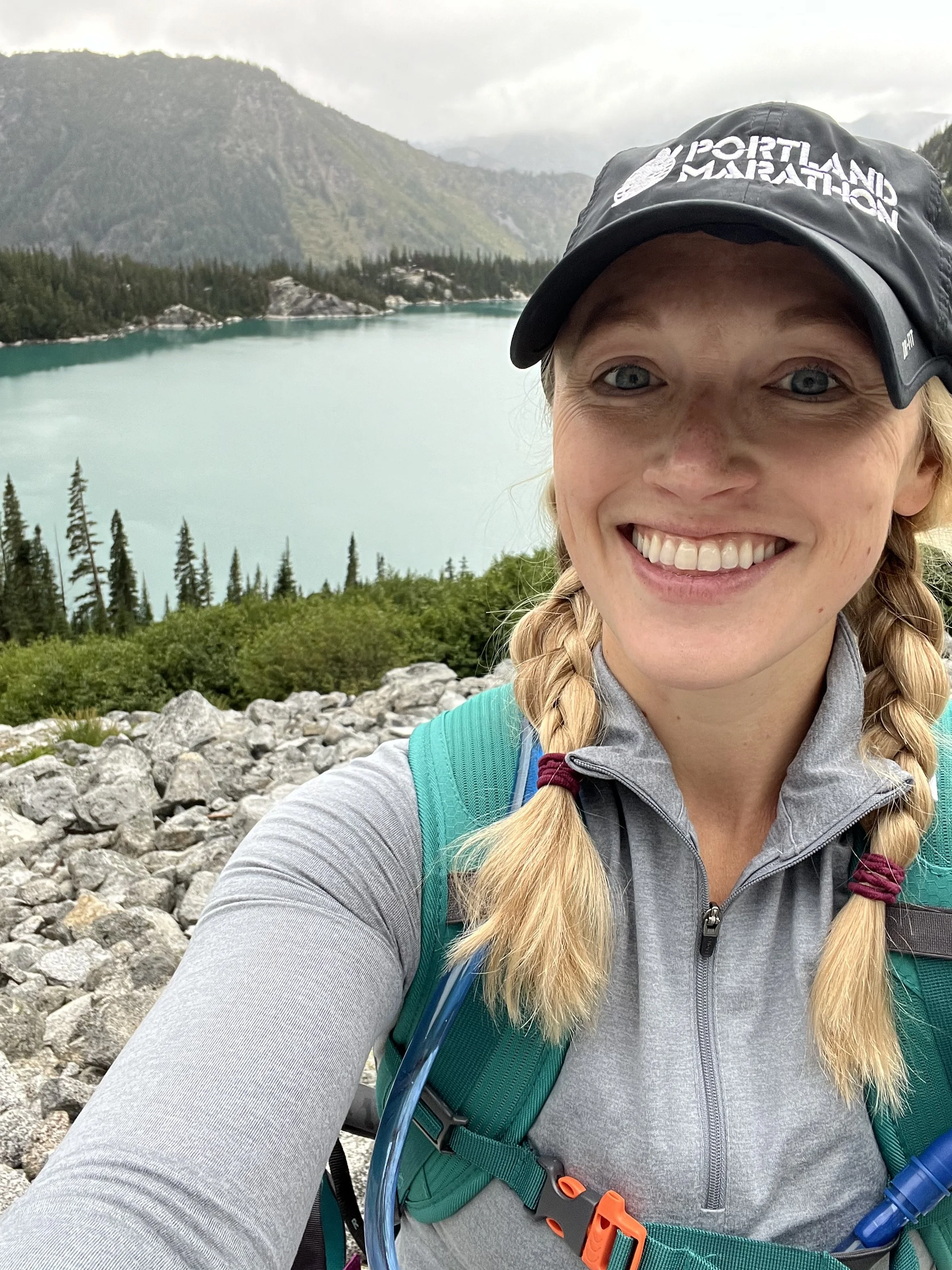 A woman hiking outdoors near a lake with mountains in the background, smiling and wearing a black Portland Marathon cap, gray jacket, and a green backpack.