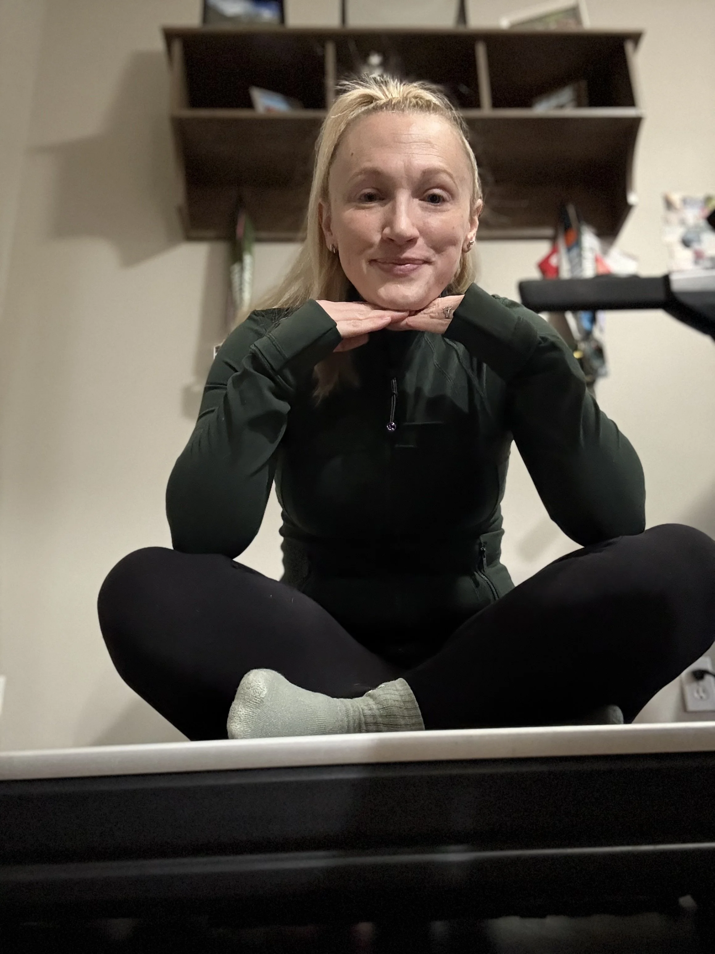 A woman sitting on the floor with her legs crossed, posing with her chin resting on her hands. She has blonde hair styled in a half-up ponytail, and is wearing a black long-sleeve athletic top and black pants. She is smiling softly and looking at the camera. Behind her, there is a wooden wall shelf with various pictures and objects, and a light-colored wall.