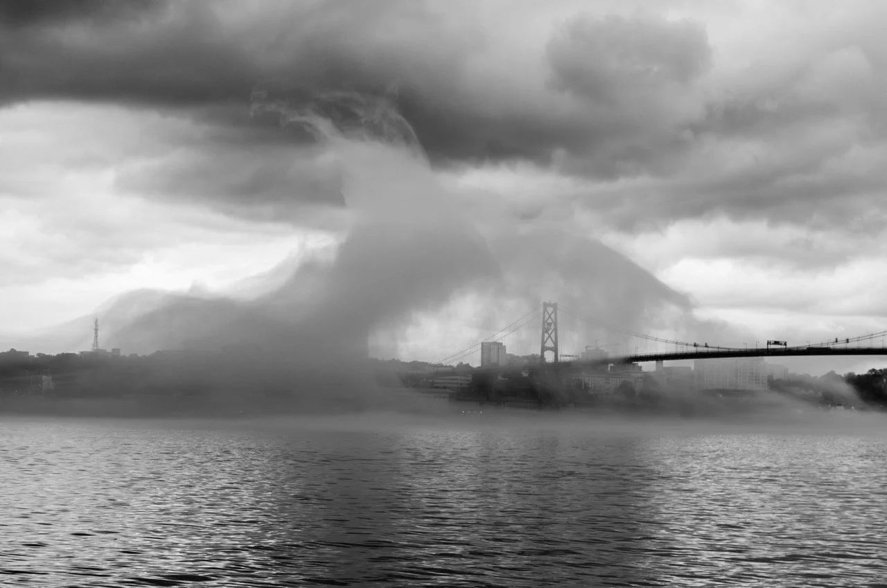 A black and white photo showing a river with fog, a suspension bridge in the background, and a large swirling cloud formation in the sky.
