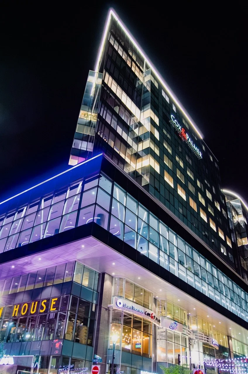A modern glass office building illuminated with colorful lights at night, displaying signs for Cox & Summer, Granville, and other businesses.