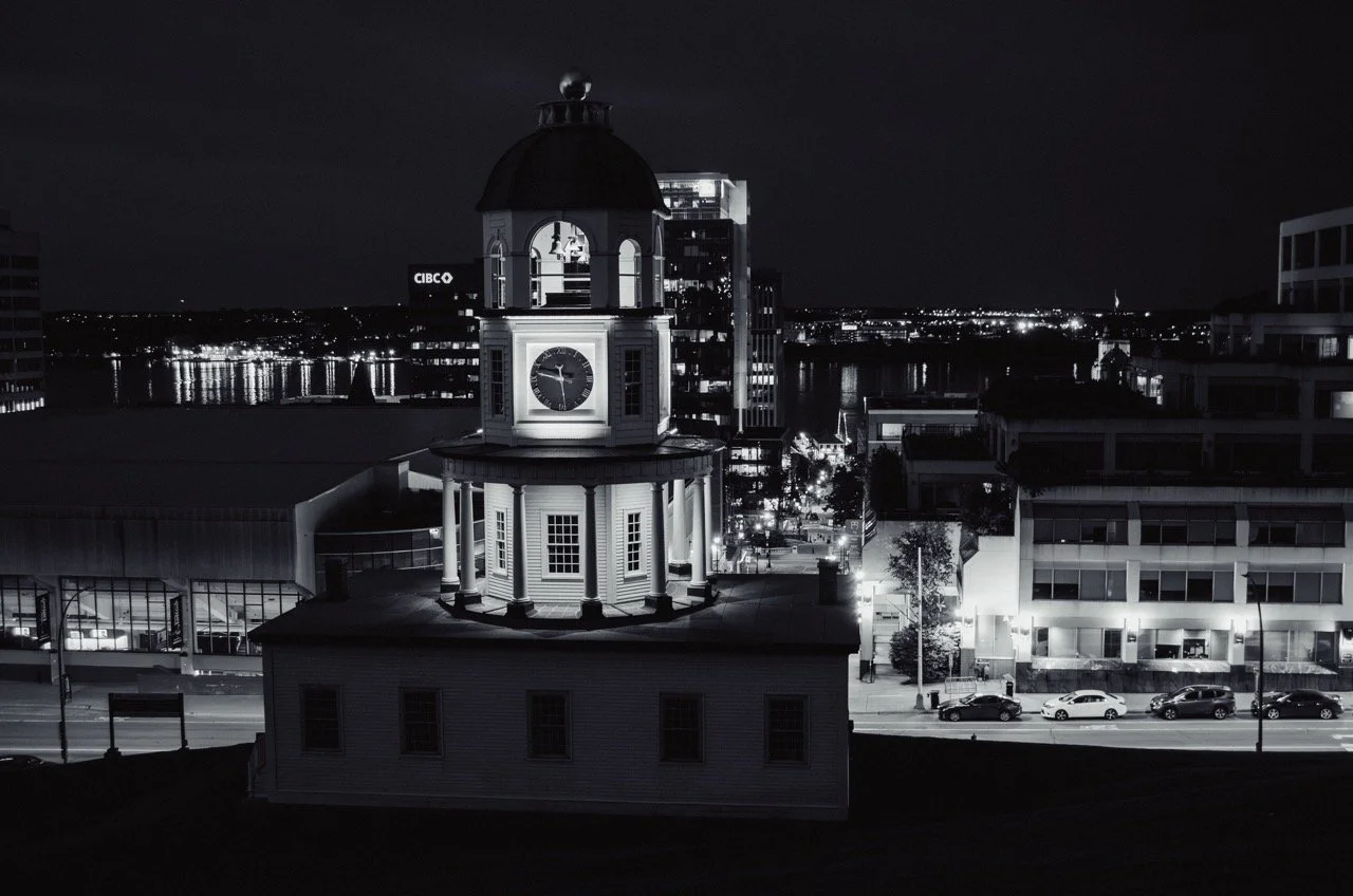 Nighttime cityscape with a historic clock tower in the foreground, illuminated against modern buildings and a river in the background, black and white image.