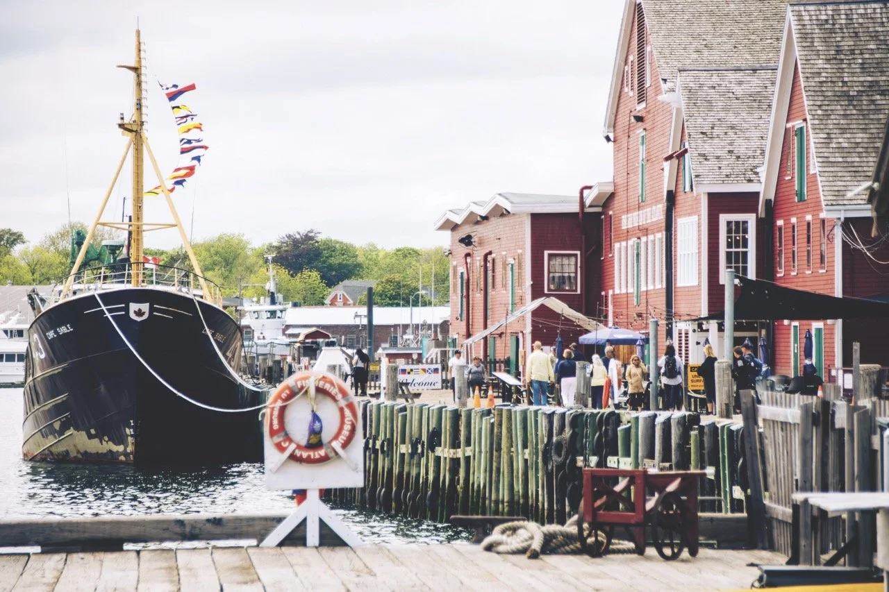 A boat docked at a harbor with red wooden buildings on the waterfront, people walking along the dock, and trees in the background.