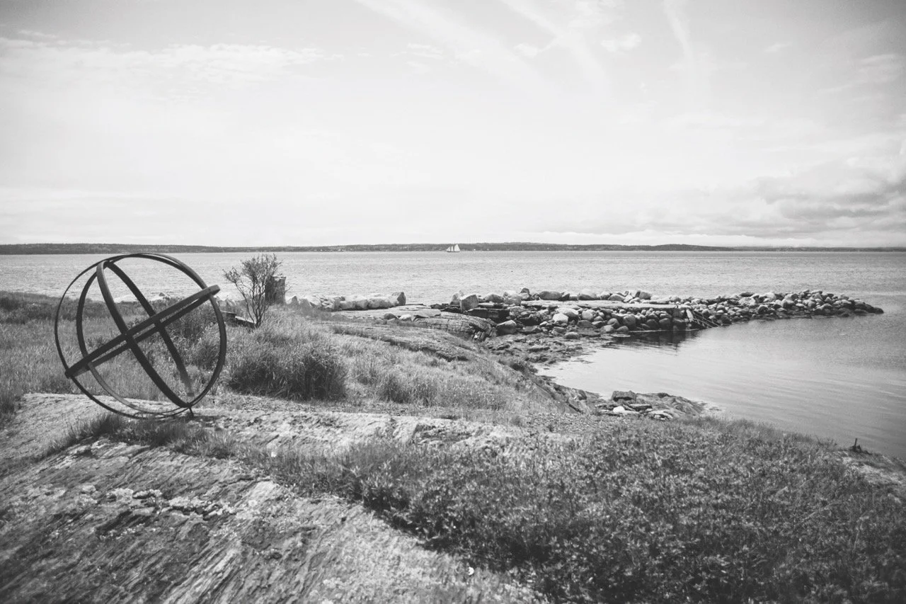 A black and white photo of a lakeside scene with a large metal spherical sculpture in the foreground, grassy terrain, rocks lining the shore, calm water, and a distant sailboat on the horizon.