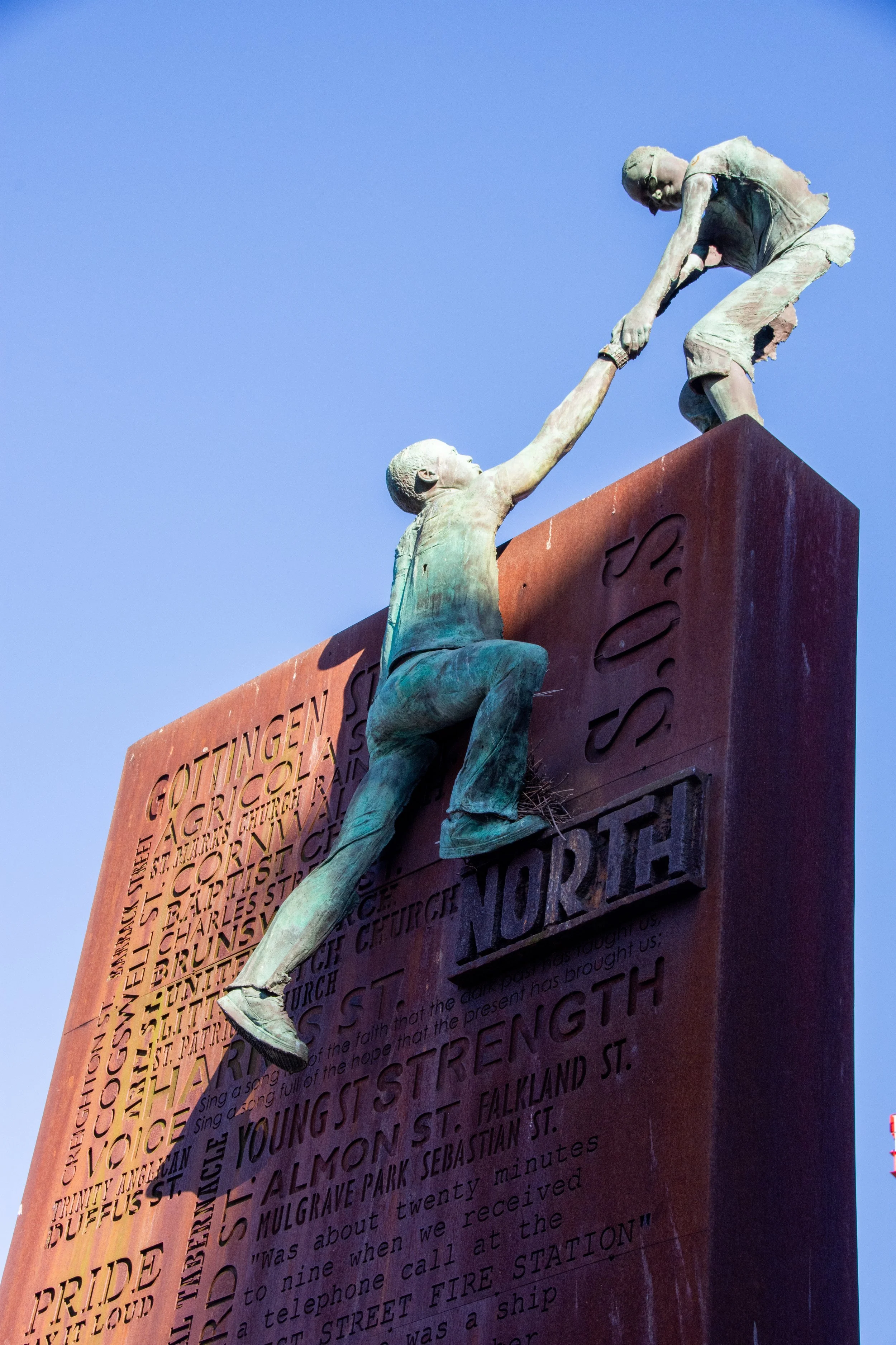 Statue of a man helping a woman climb a wall with engraved words and quotes, set against a clear blue sky.