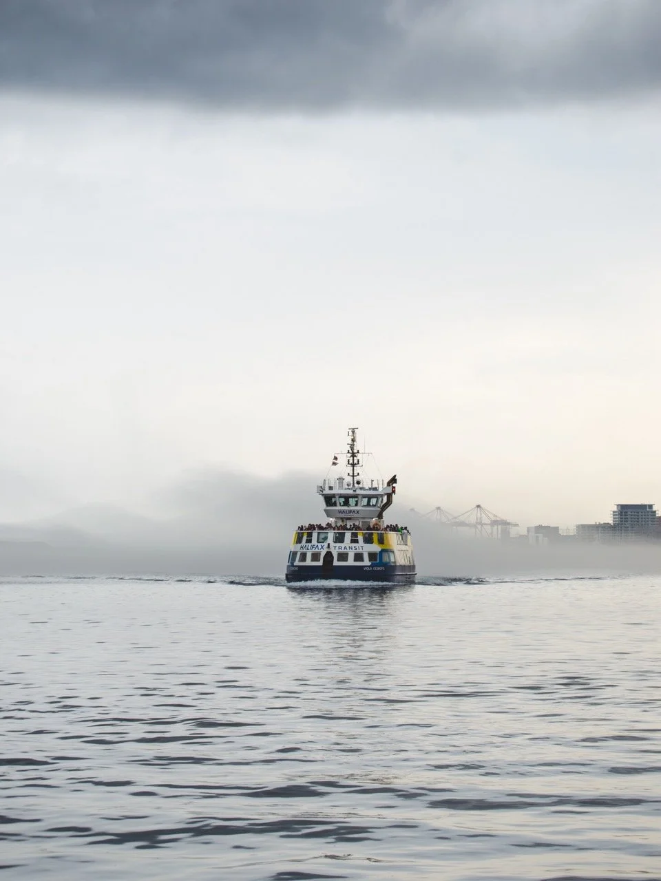 A ferry boat labeled 'Halifax Transit' navigating through misty waters with a city skyline and cranes in the background.