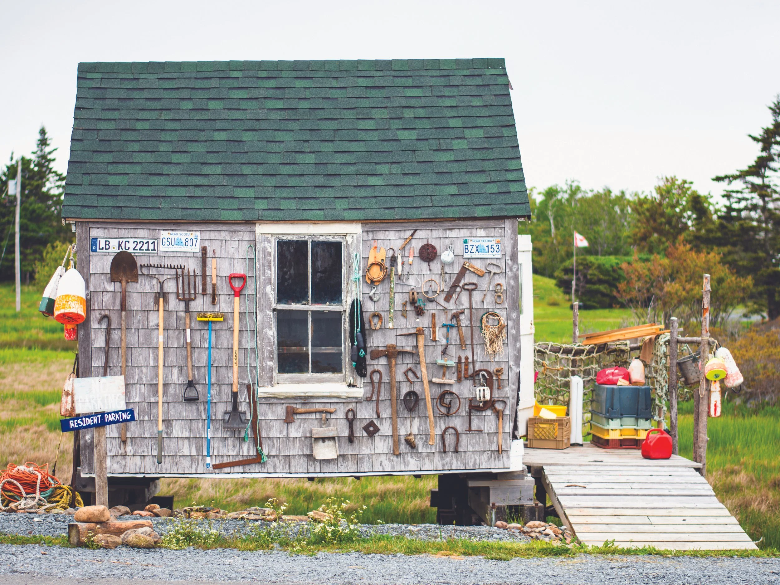 A small wooden shed with gardening tools hung on the exterior wall near a window, a ramp leading up to the shed, and a sign indicating vehicle parking for residents, set in a rural area with trees and grass in the background.