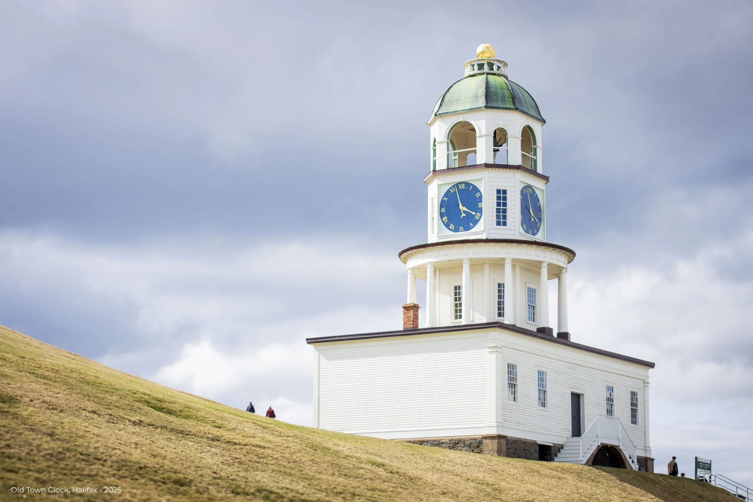 A historic white building with a clock tower on top, featuring two large blue clock faces showing approximately 1:55, set on a grassy hill with a cloudy sky in the background.