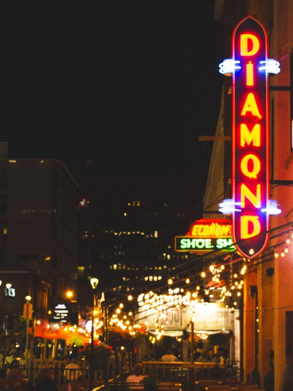 Night view of a city street with illuminated neon signs, including a large vertical 'DIA MOND' sign and string lights hanging above outdoor dining area.