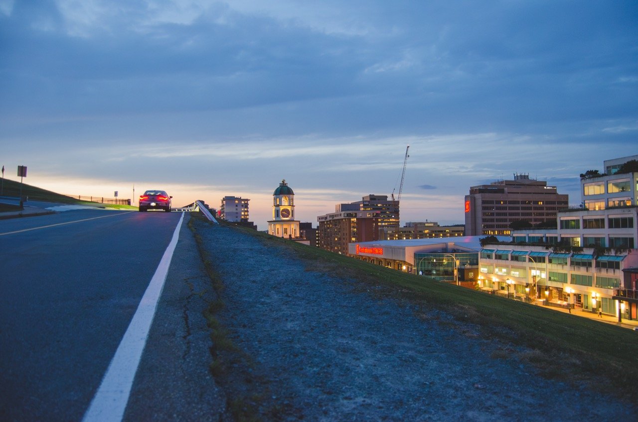City skyline at dusk with buildings, a clock tower, and a car on the road in the foreground.