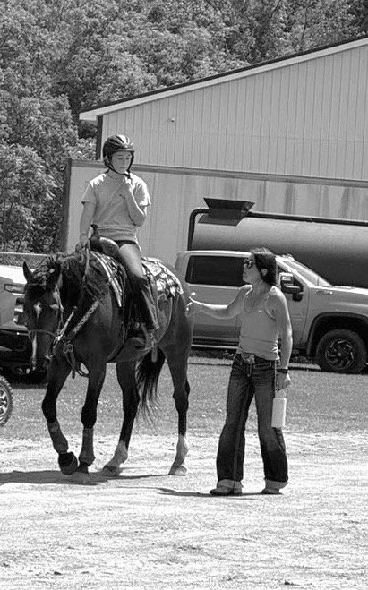 A girl riding a horse with a person beside her, holding the horse's reins, in an outdoor area with a building and vehicles in the background.