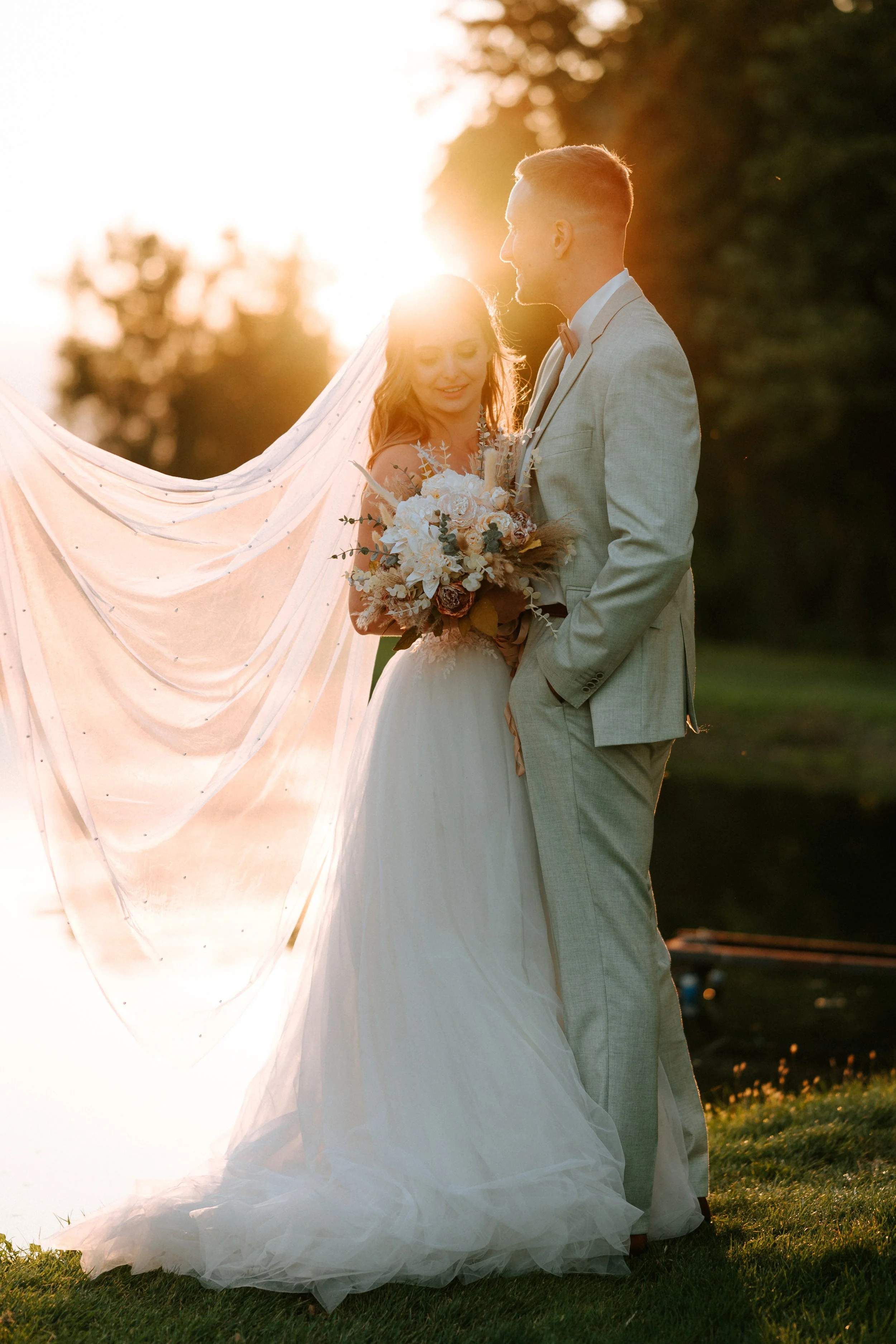 A bride and groom standing outdoors at sunset, with the bride holding a bouquet of flowers and the groom wearing a light gray suit with a bow tie, sharing a moment together.