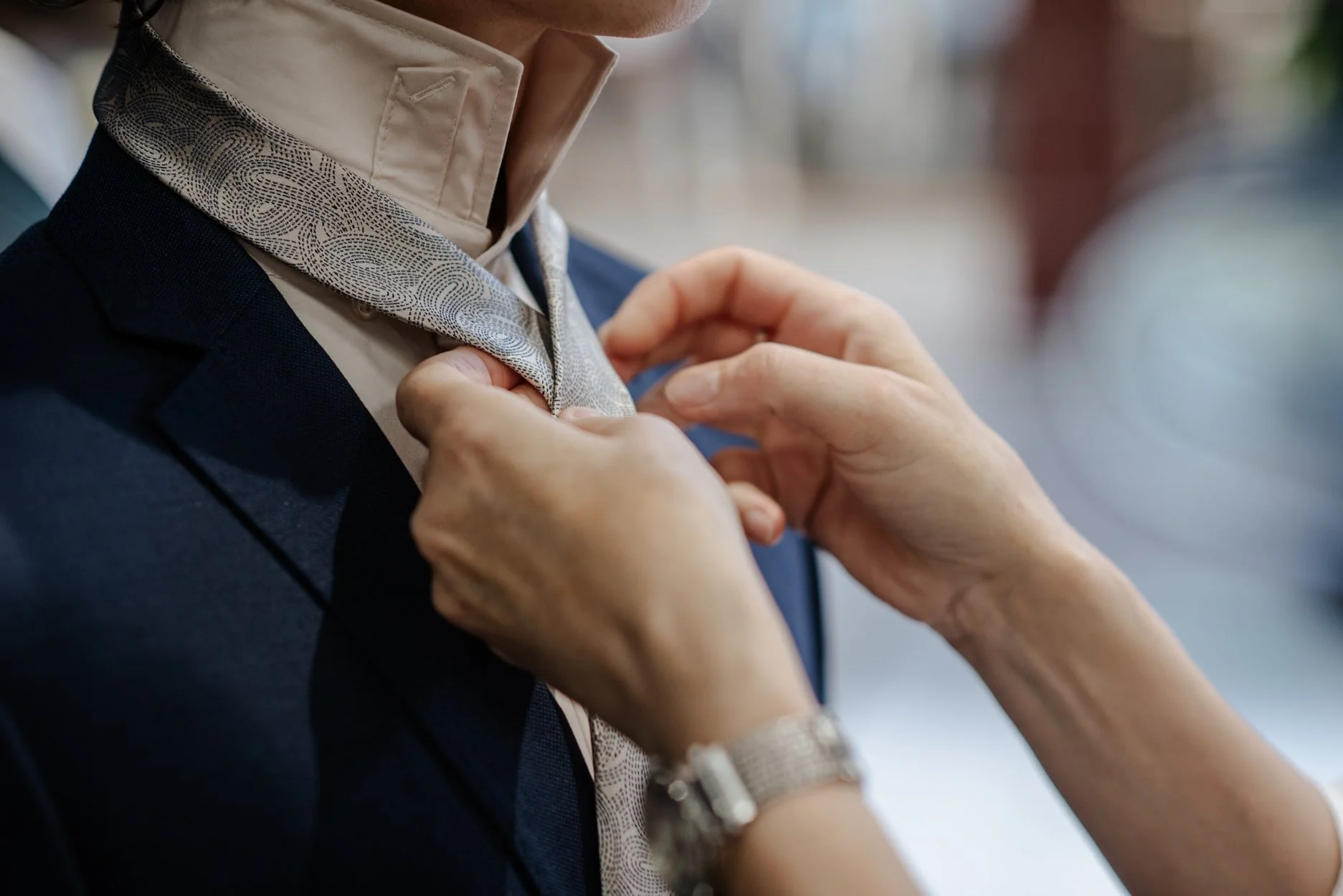 A person is adjusting their collared shirt while wearing a dark blazer. The focus is on their hands and the upper part of their body, with a patterned scarf or tie being adjusted around their neck.