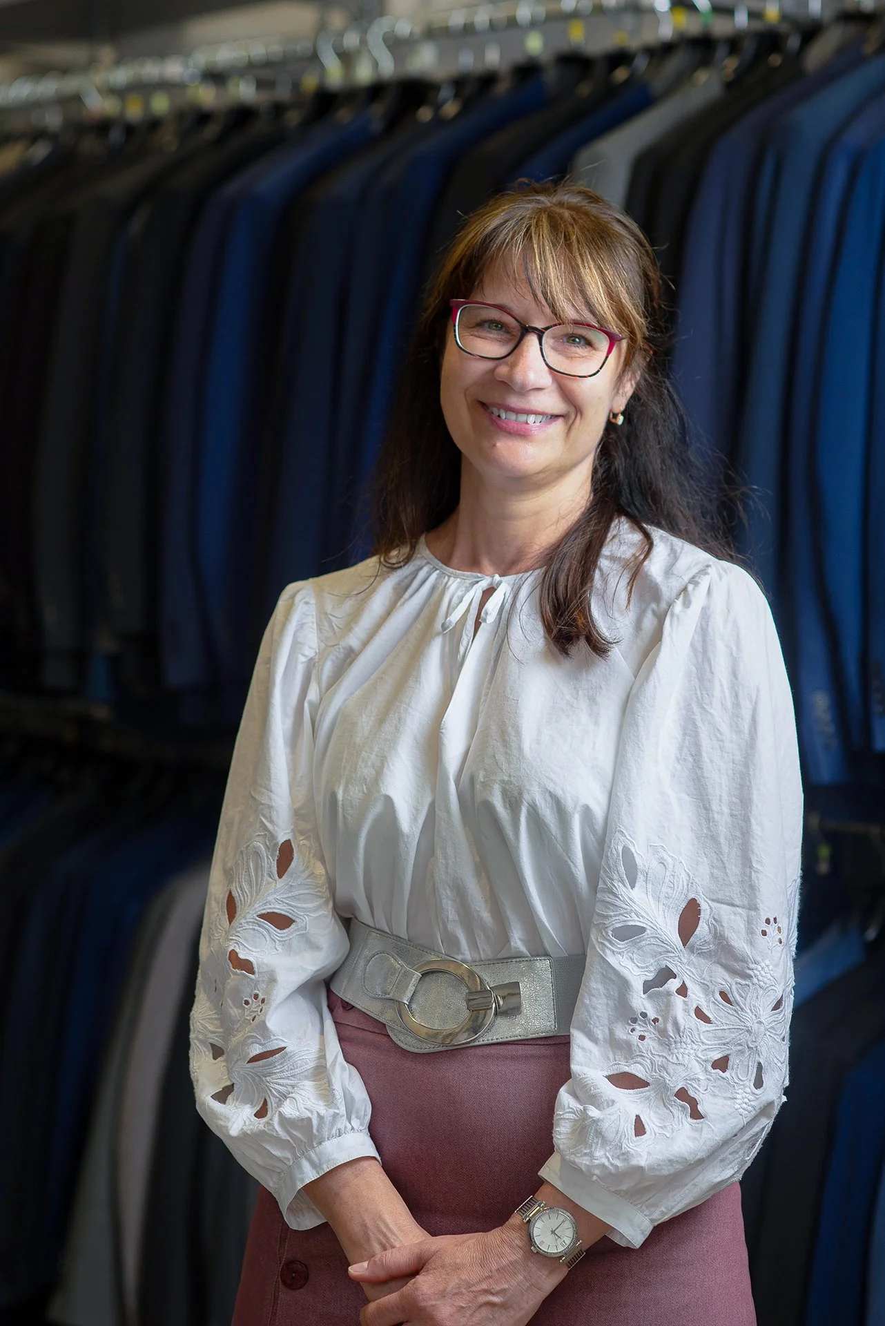 A woman wearing glasses and a white blouse with eyelet details stands in front of a row of hanging suits or blazers in a clothing store.