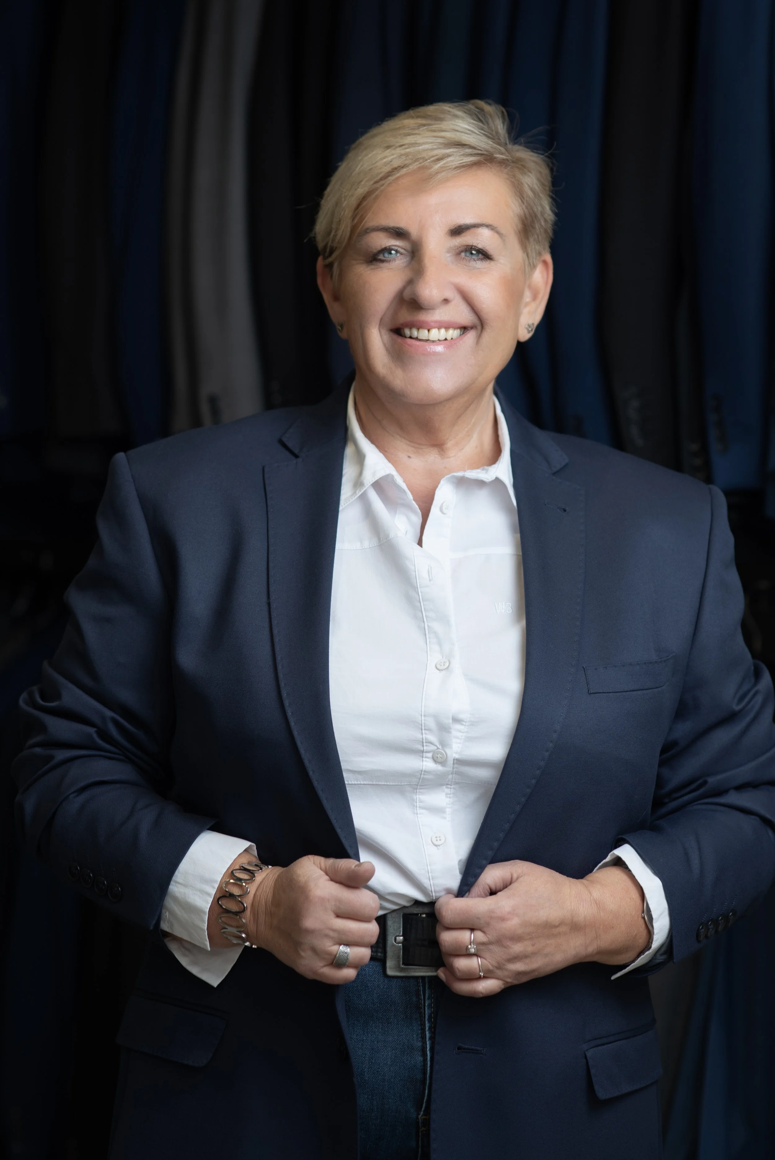 A woman with short blonde hair, blue eyes, and a bright smile, wearing a dark blue blazer and a white button-up shirt, standing against a backdrop of hanging dark clothing, with her hands clasped in front of her.