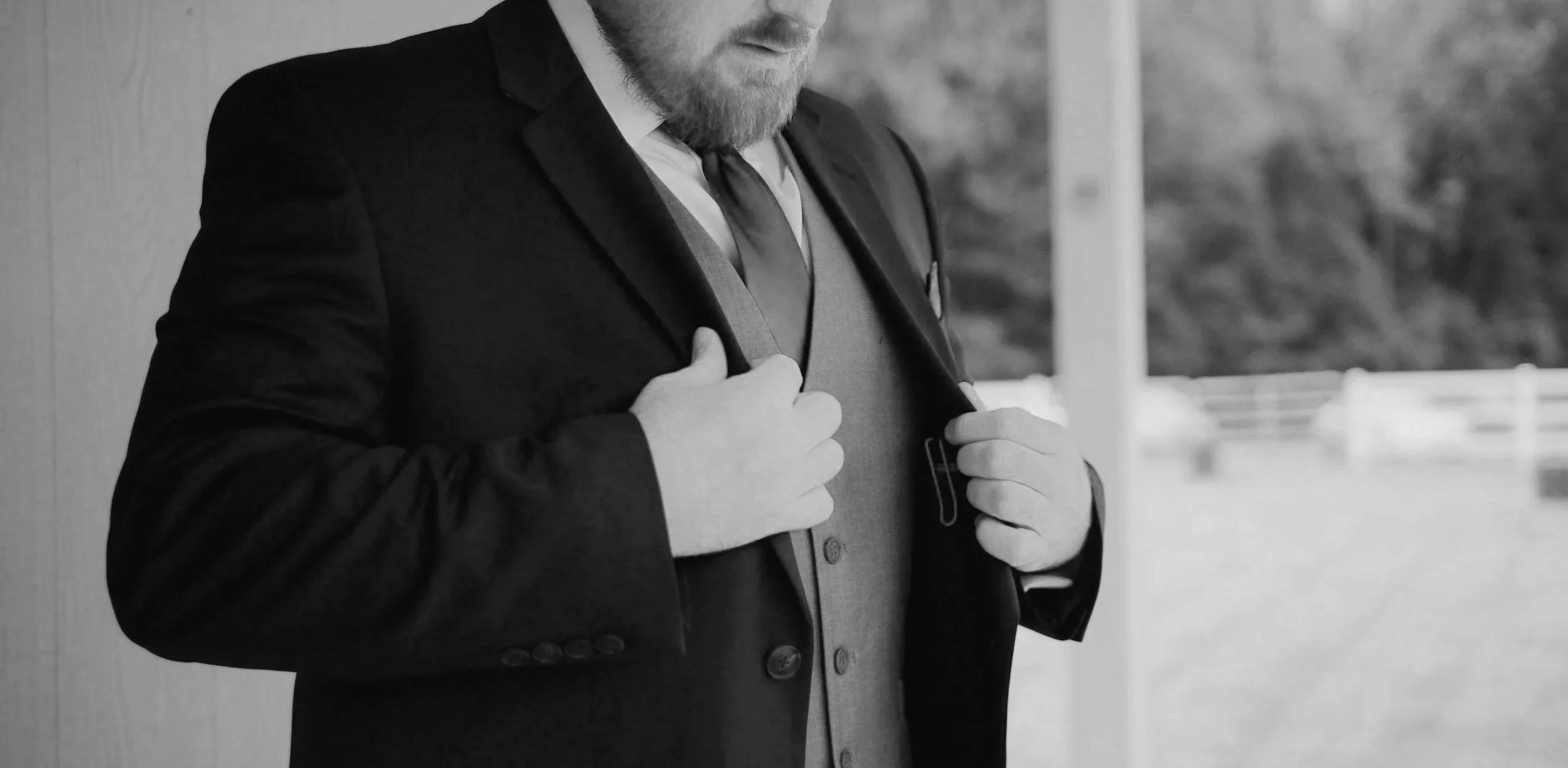 A man in a suit adjusting his jacket, with a backpack strap over his shoulder, standing outdoors near a wall.