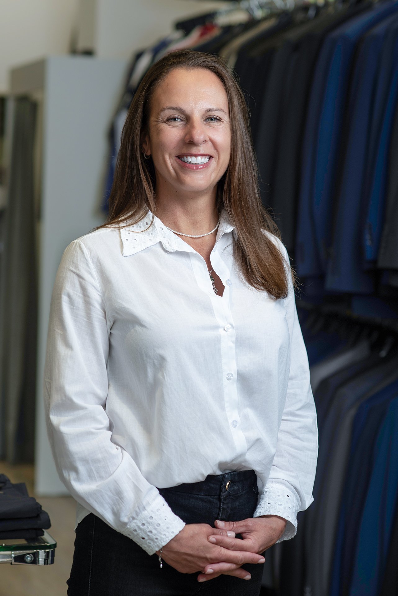 A woman with long brown hair, smiling, wearing a white button-up shirt with lace details on the collar and cuffs, standing in front of clothing racks filled with suits and dress shirts in a retail store.