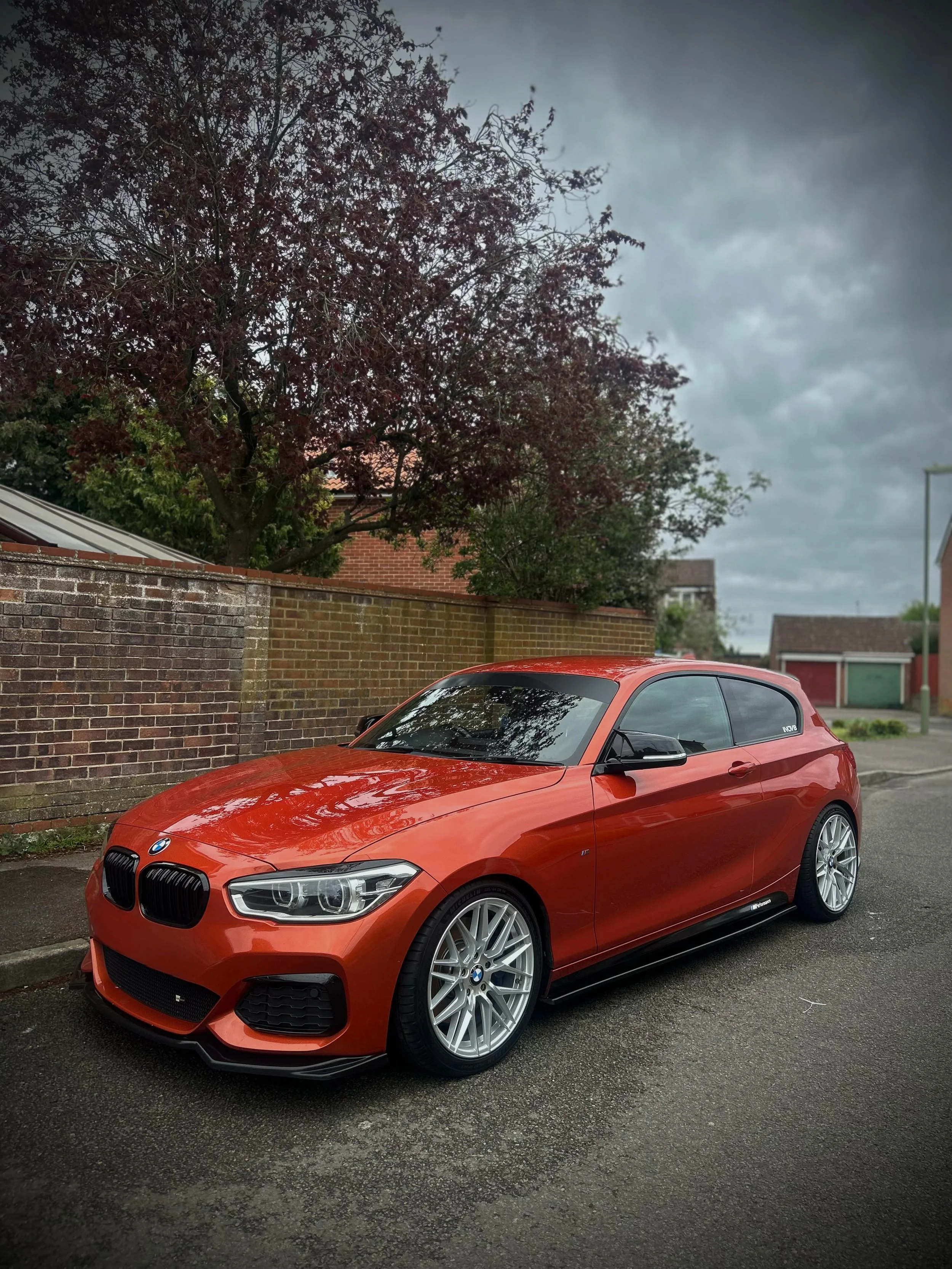 A bright red BMW car parked on the street in front of a brick wall and a tree with dark red leaves on a cloudy day.