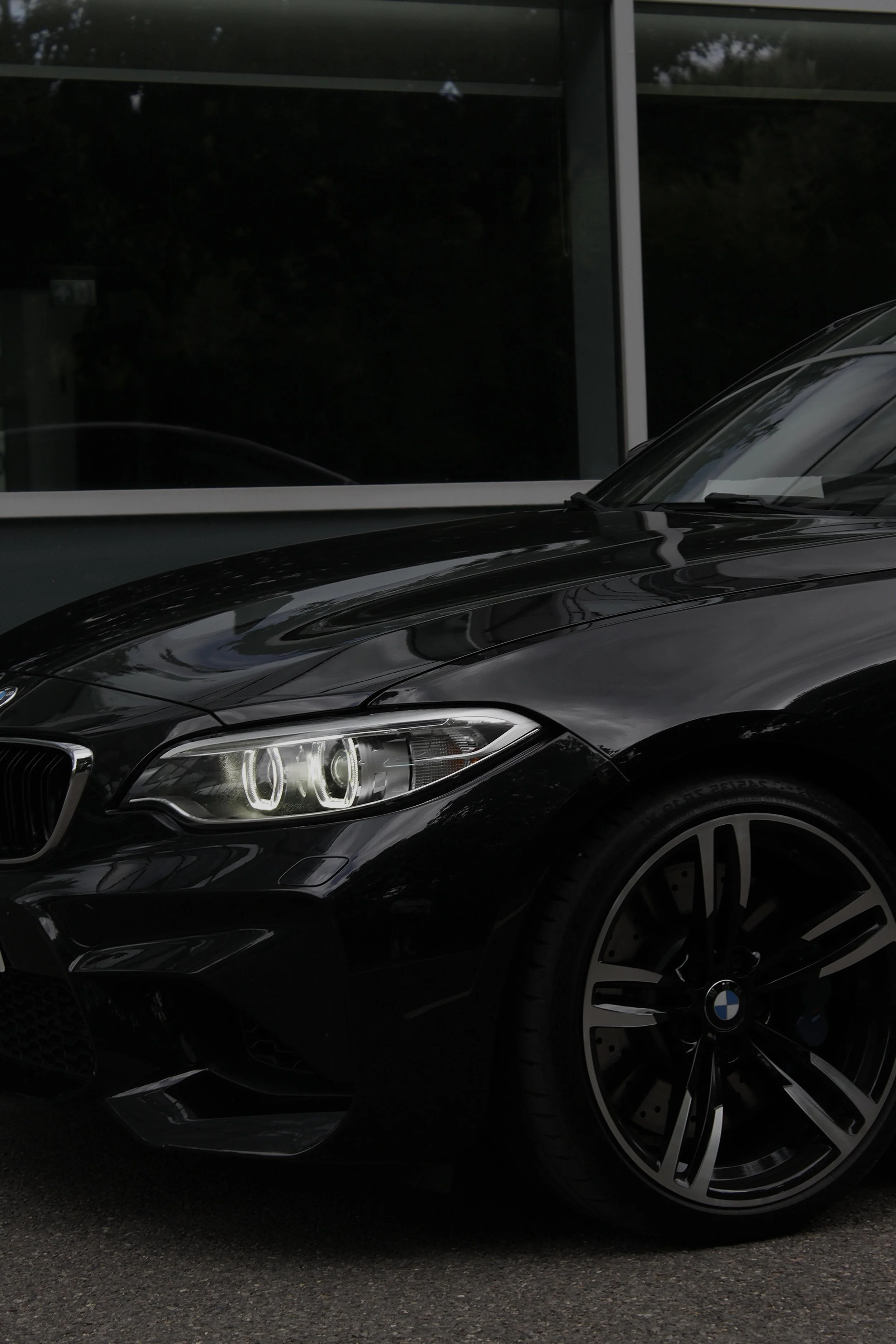 Close-up of a black BMW car with illuminated headlights, parked on a road in front of a modern building with large reflective windows.