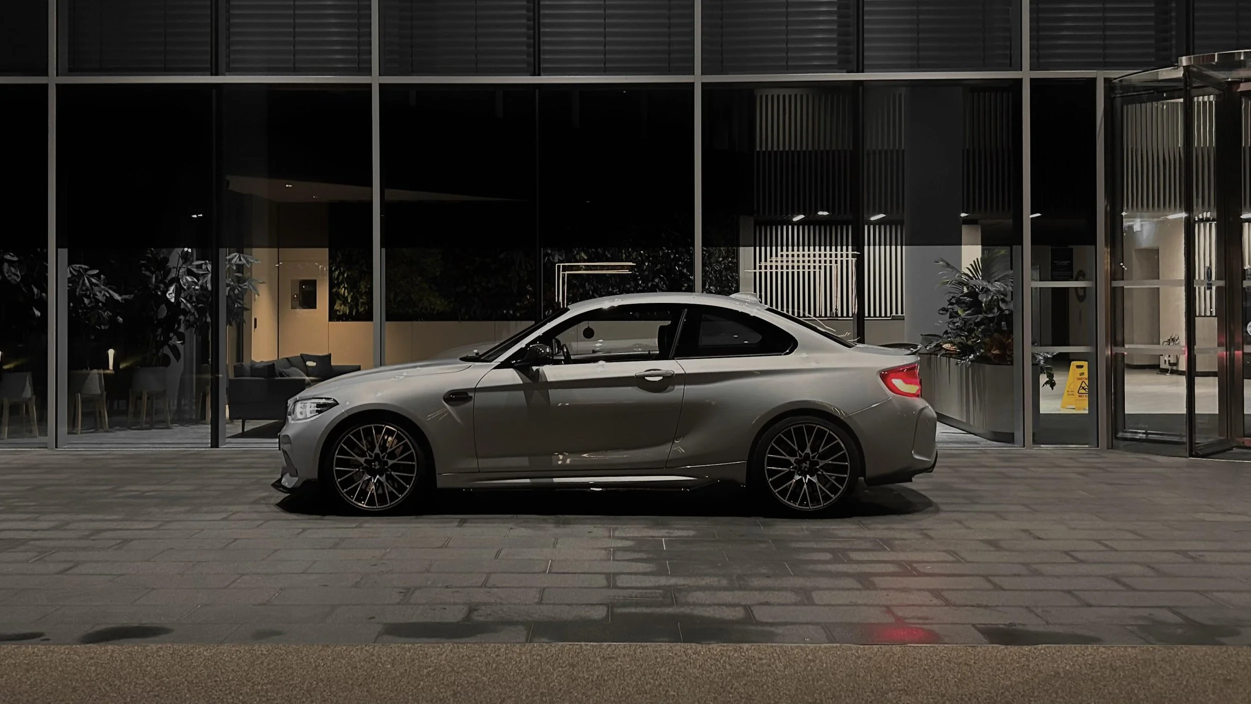 A silver BMW car parked outside a modern building with glass windows and a seating area visible inside.