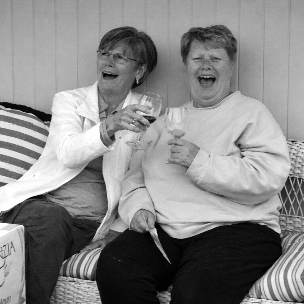 Two women sitting on a striped cushion, holding glasses of wine, laughing and enjoying each other's company.