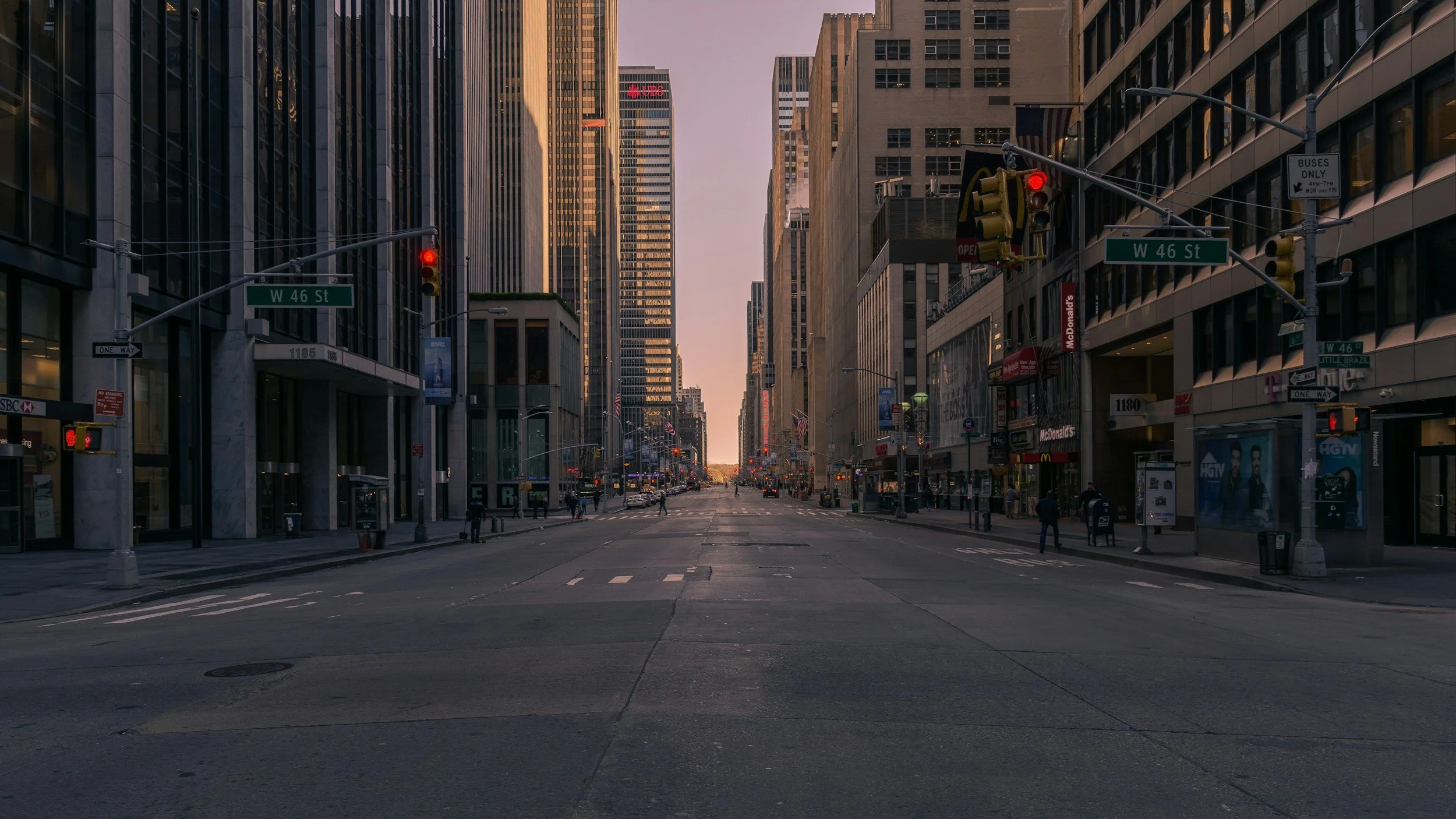 Empty city street at dawn with tall buildings on each side, traffic lights, and a few pedestrians walking, view down a long avenue.