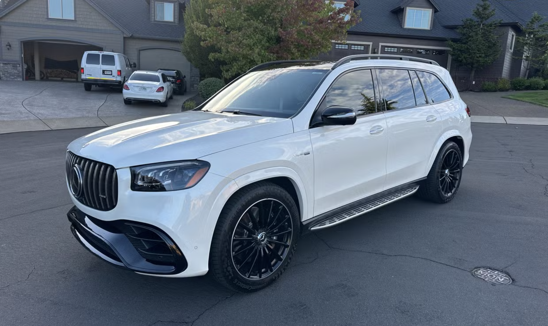 White luxury SUV parked on a driveway in front of a suburban house with other vehicles and trees in the background.