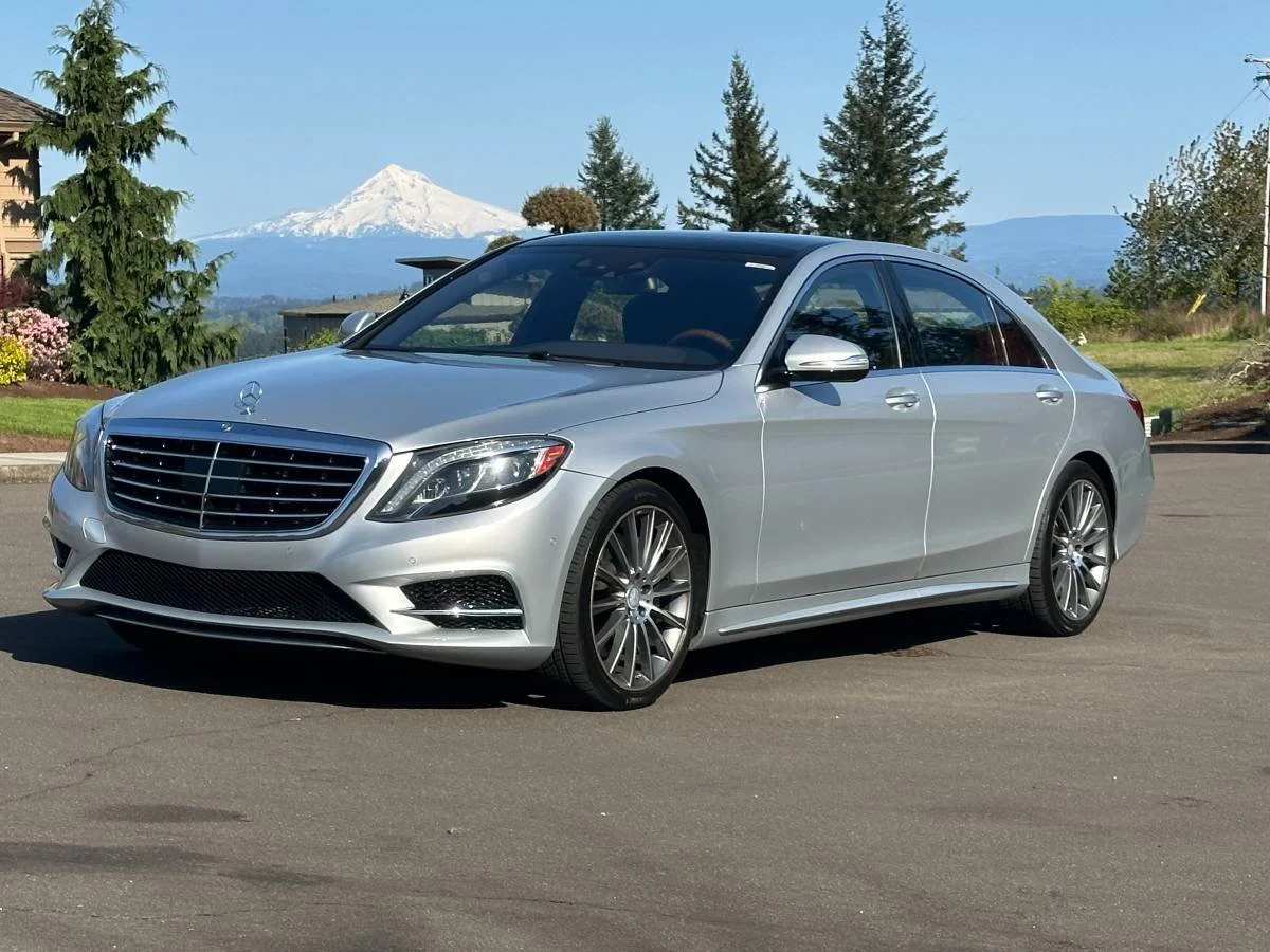 A silver Mercedes-Benz sedan parked on a paved road with a backdrop of trees, a house, and Mount Rainier in the distance.