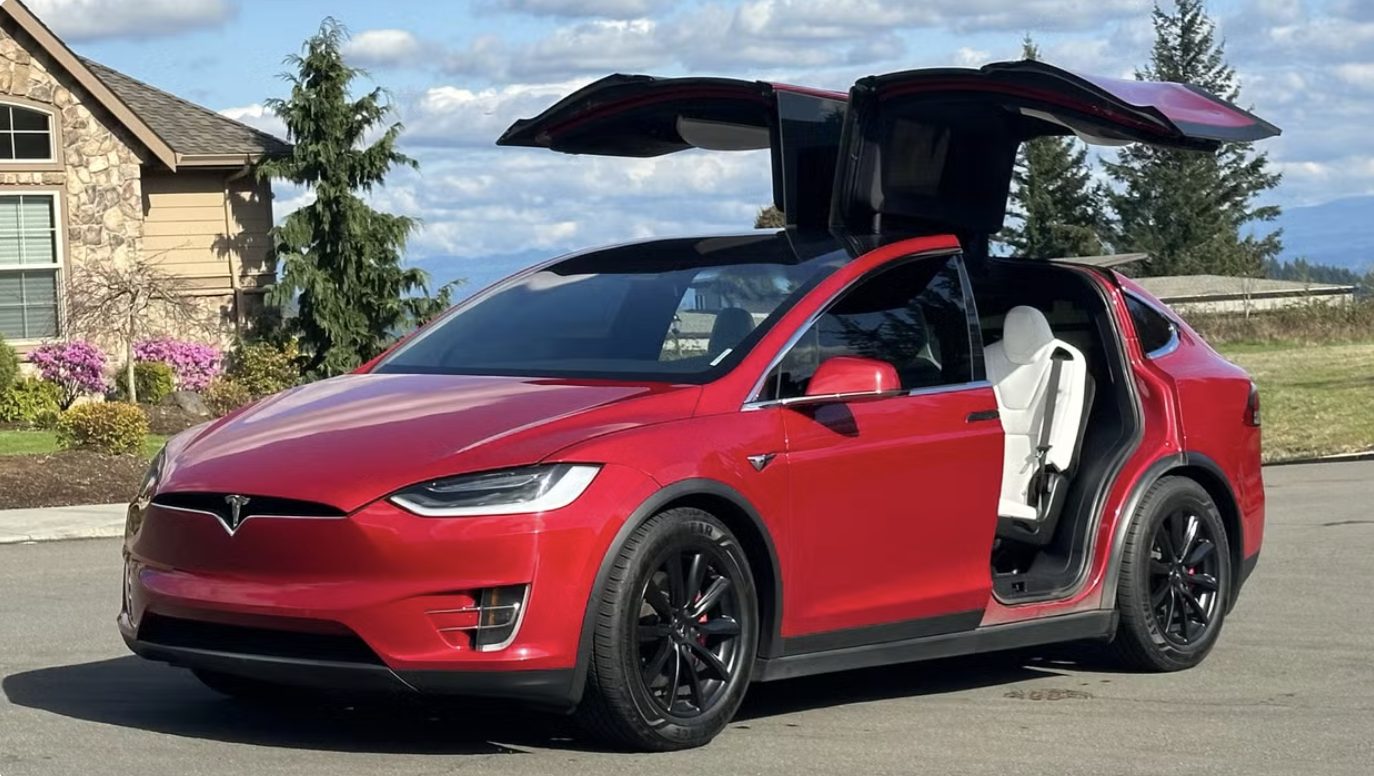 Red Tesla Model Y with falcon-wing doors open, parked on a suburban street with a house and trees in the background.