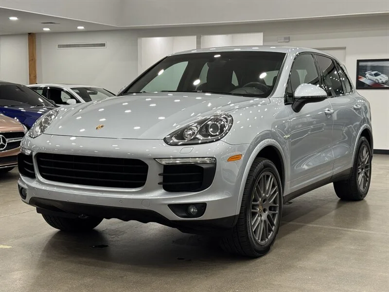 A white Porsche SUV parked indoors at a car dealership, with other vehicles visible in the background.