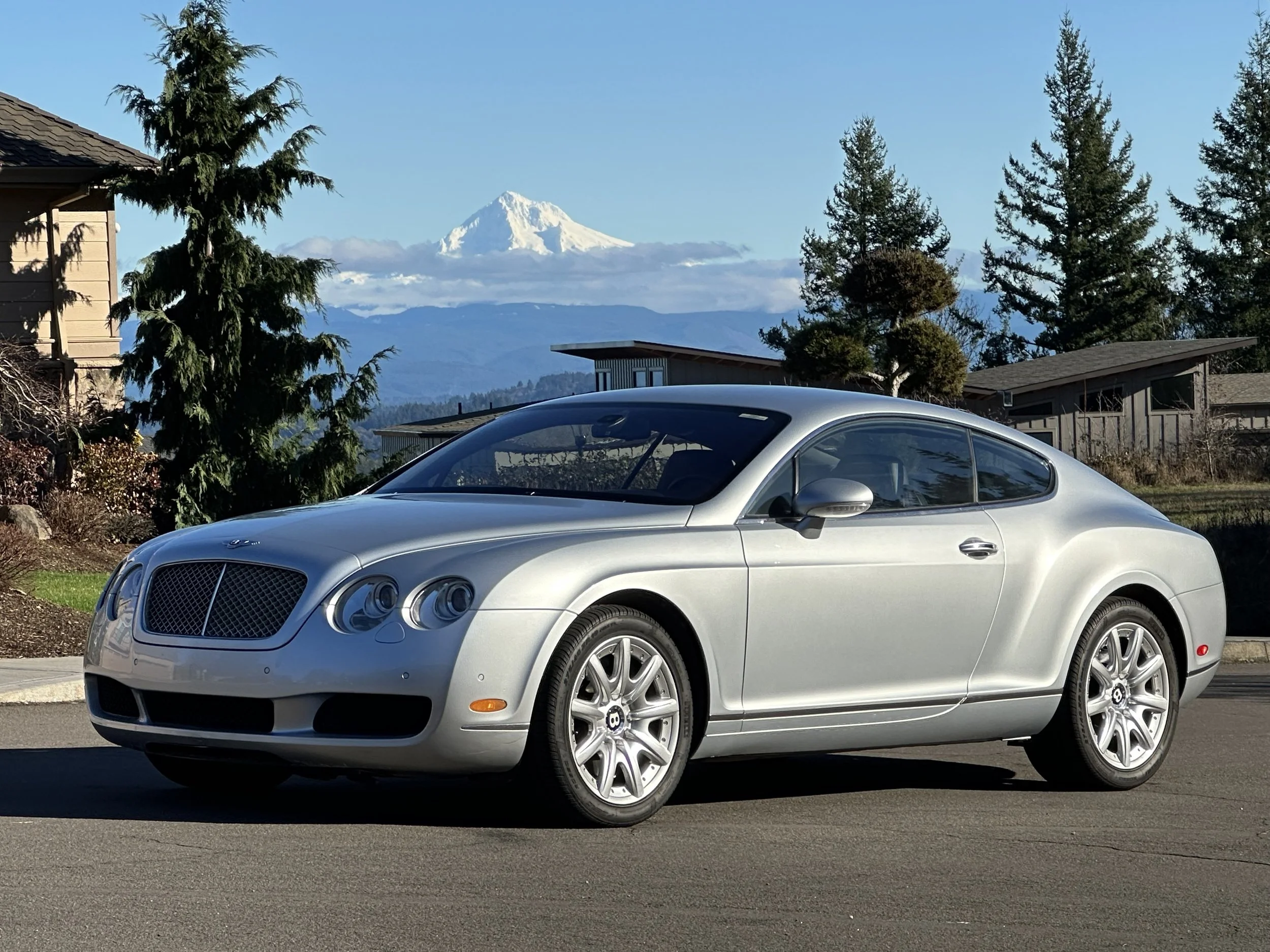 A silver Bentley coupe parked on a suburban street with trees, houses, and Mount Rainier in the background.