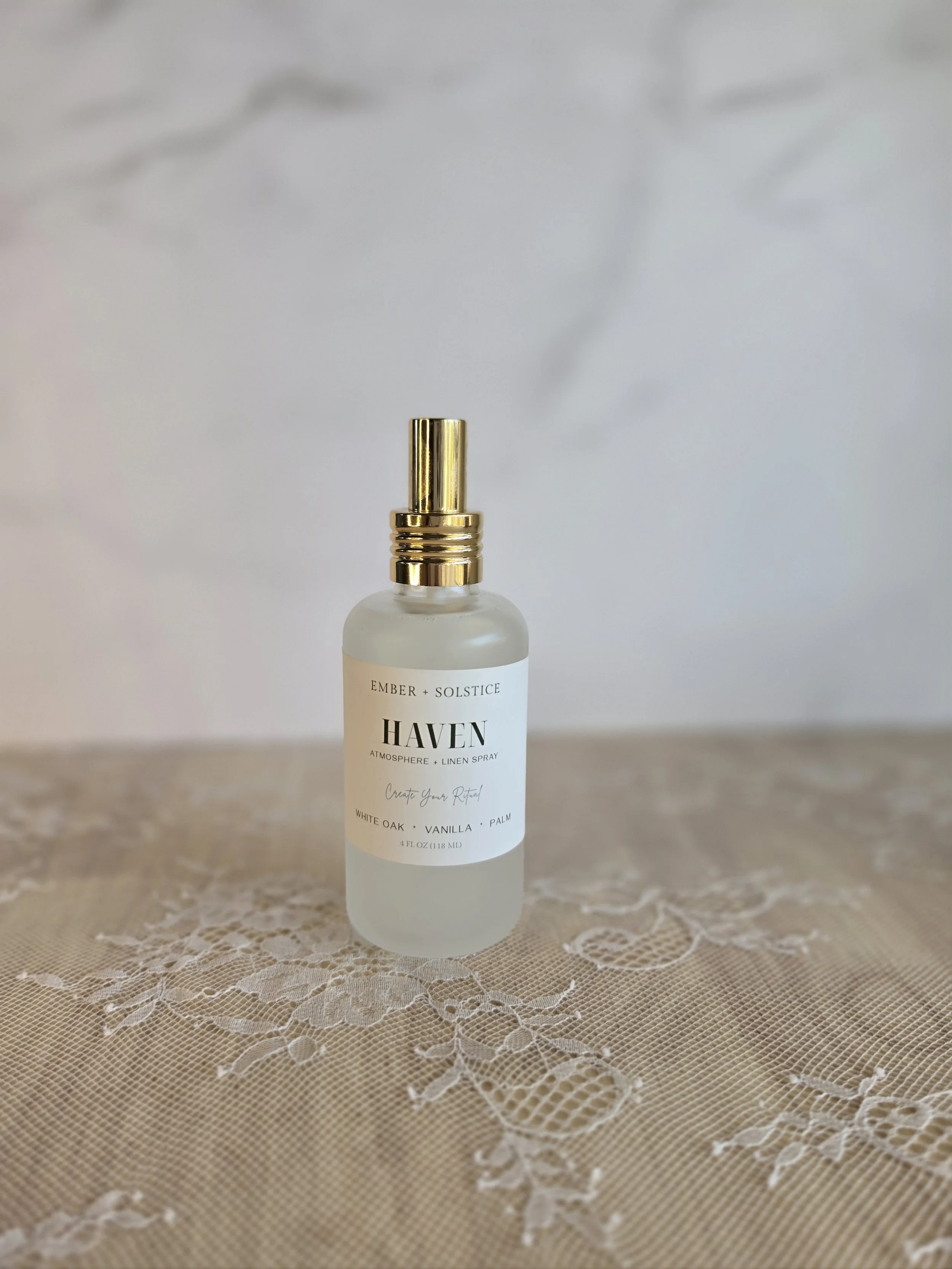 A frosted glass spray bottle with a gold spray top labeled 'Haven Atmosphere + Linen Spray' rests on a lace tablecloth.