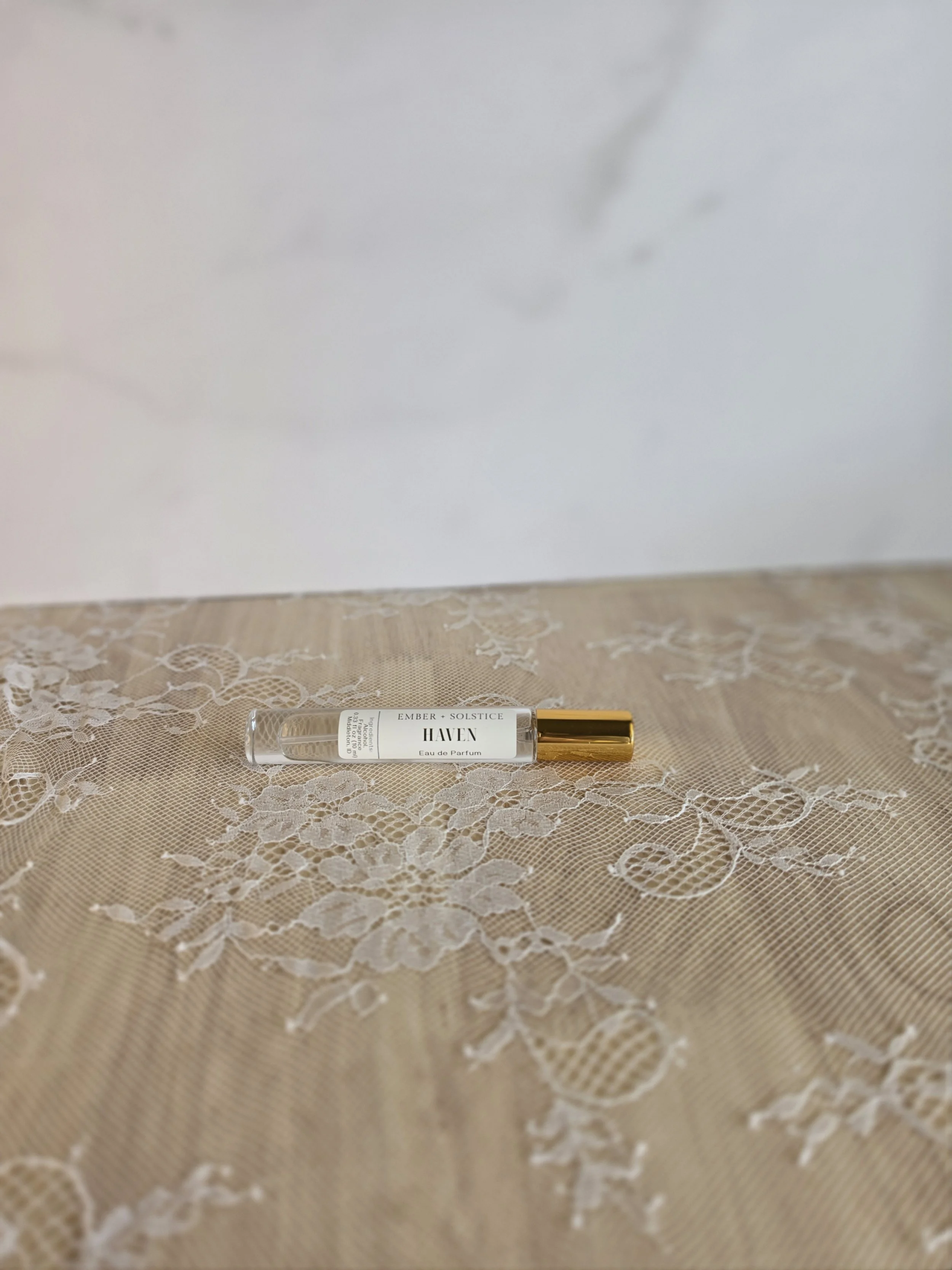 A bottle of perfume labeled 'HAVEN' with a gold cap, placed on cream-colored lace fabric, with a plain white wall background.