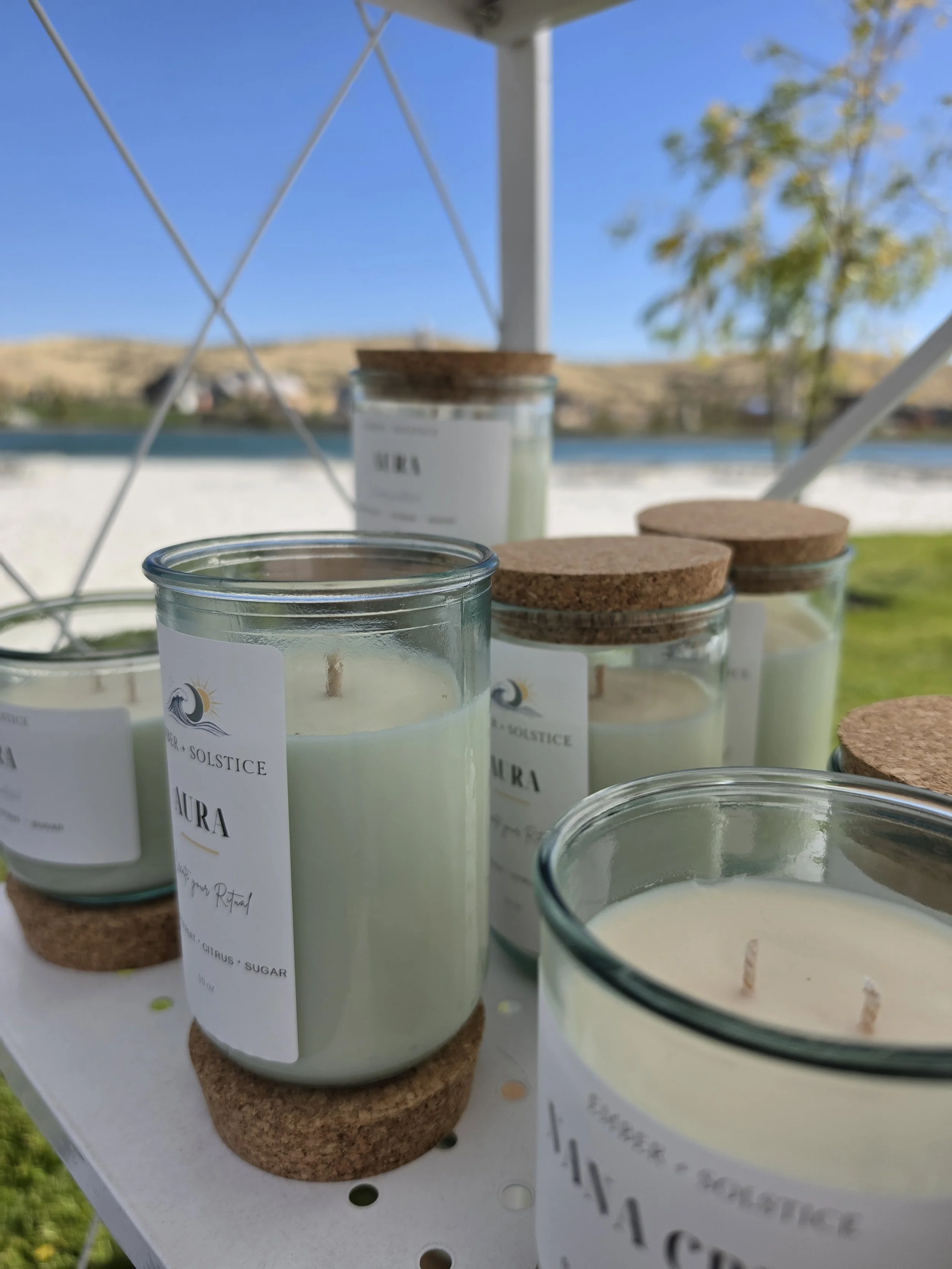 Several white candles in glass jars with cork lids displayed on a white surface outdoors with a lake and trees in the background.