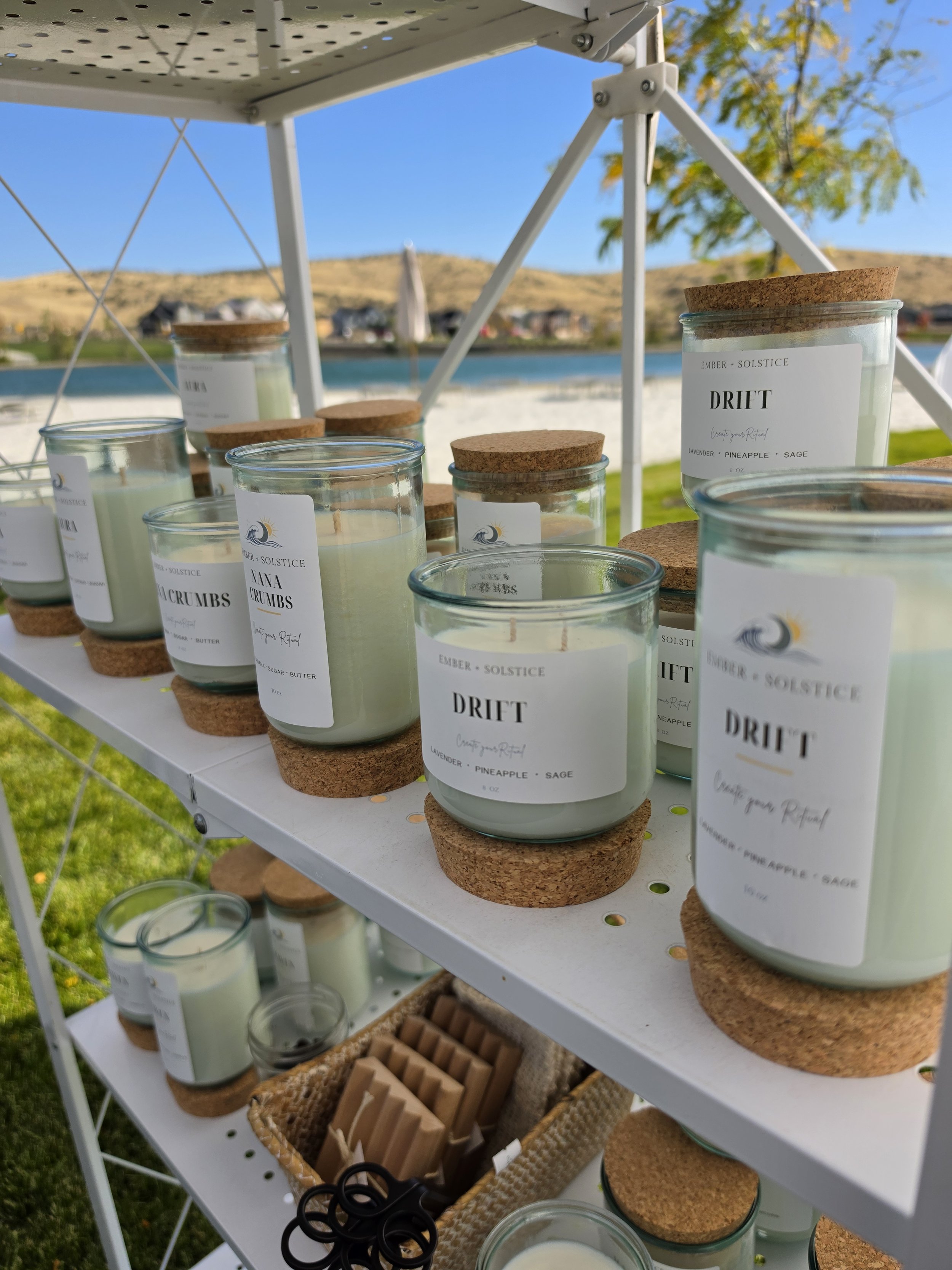 Outdoor display of scented candles in glass jars with cork lids on a white metal shelf with wooden coasters underneath, set against a scenic background of a lake, trees, and hills under a blue sky.