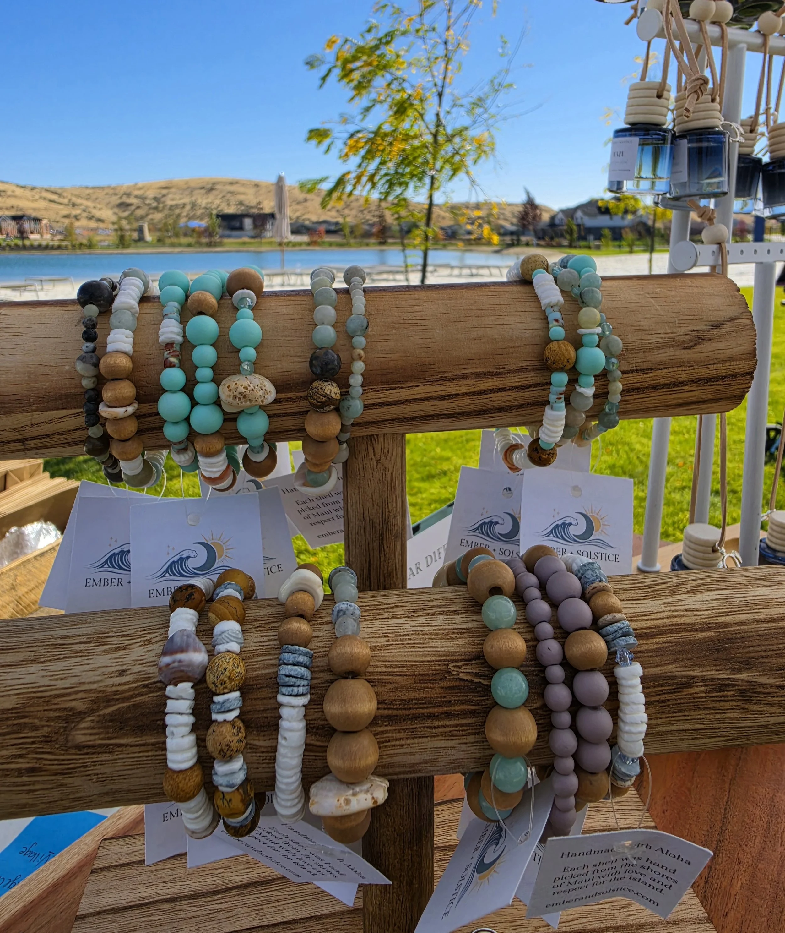 Display of handcrafted beaded jewelry including necklaces, hanging from a wooden stand outdoors with a lake, trees, and hills in the background under a blue sky.