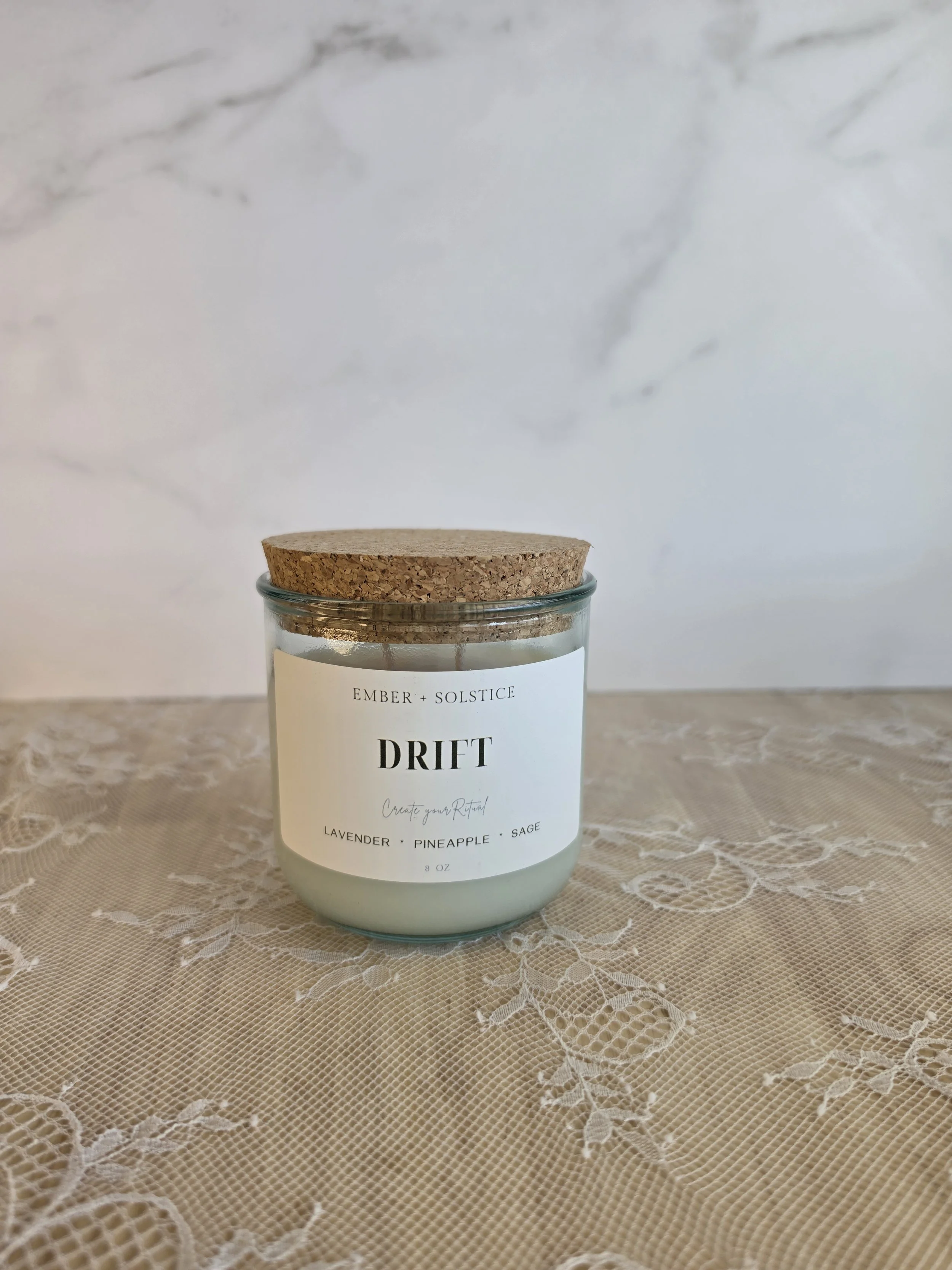 A white scented candle in a glass jar with a cork lid, labeled 'DRIFT' by Ember + Solstice, placed on a lace tablecloth with a marble wall background.