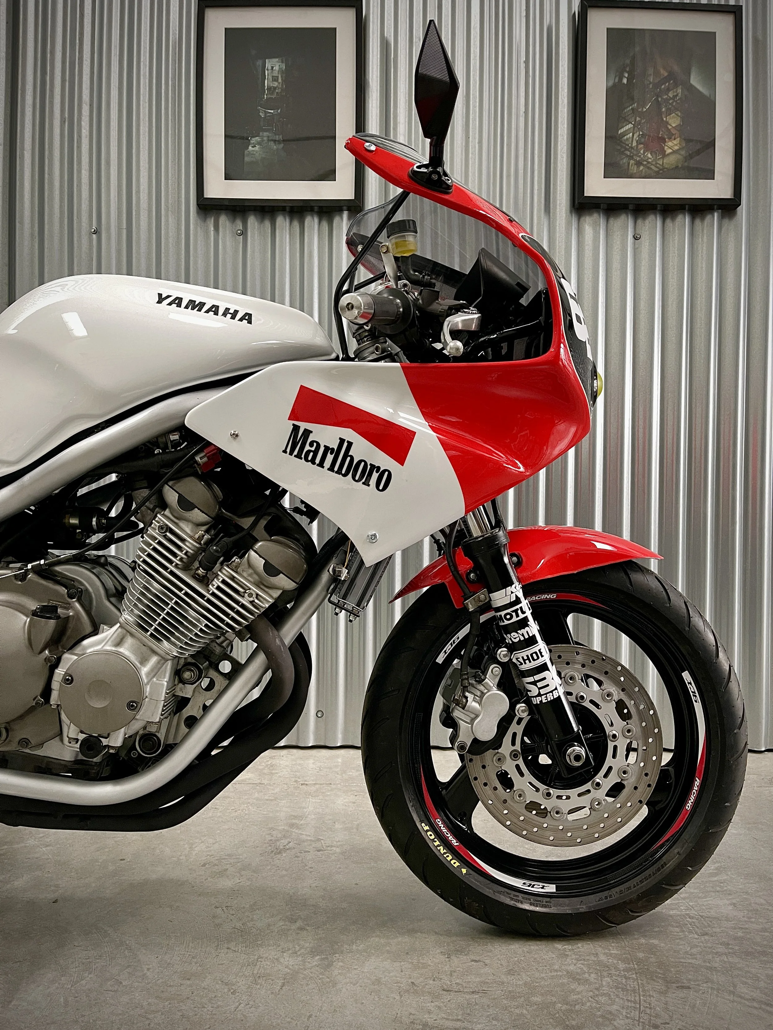 Yamaha XJ600 Diversion 1992, transformada estilo endurance años 80, en un ambiente de taller o exhibición, con paredes de metal y dos cuadros en el fondo.