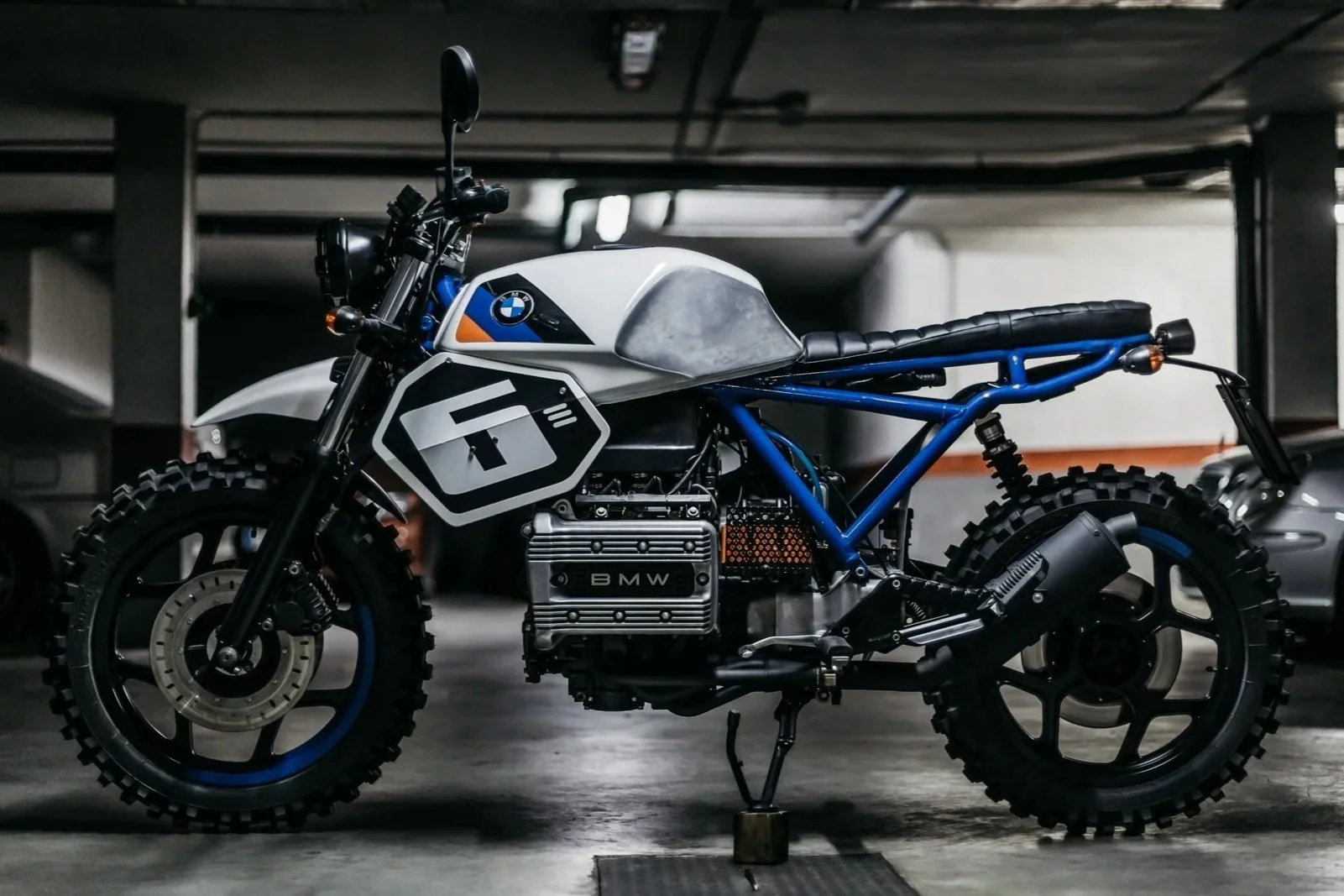 Motocicleta en un estacionamiento subterráneo, con diseño de estilo retro y elementos modernos, marca BMW, con estructura azul y detalles en negro, sin carrocería convencional.